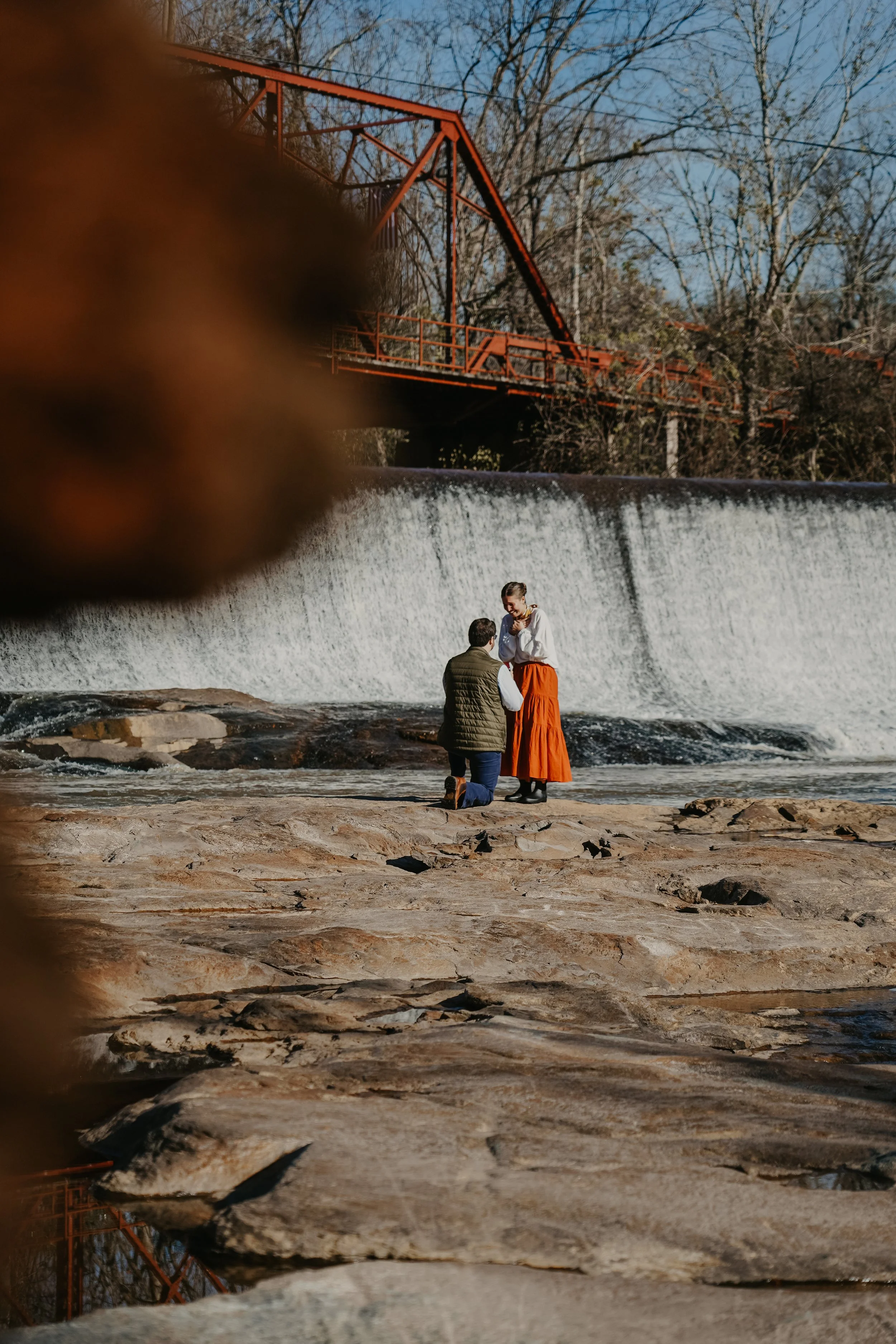 A couple kneeling on rocks by a waterfall, with a woman smiling and touching her face, while the man holds her hands.