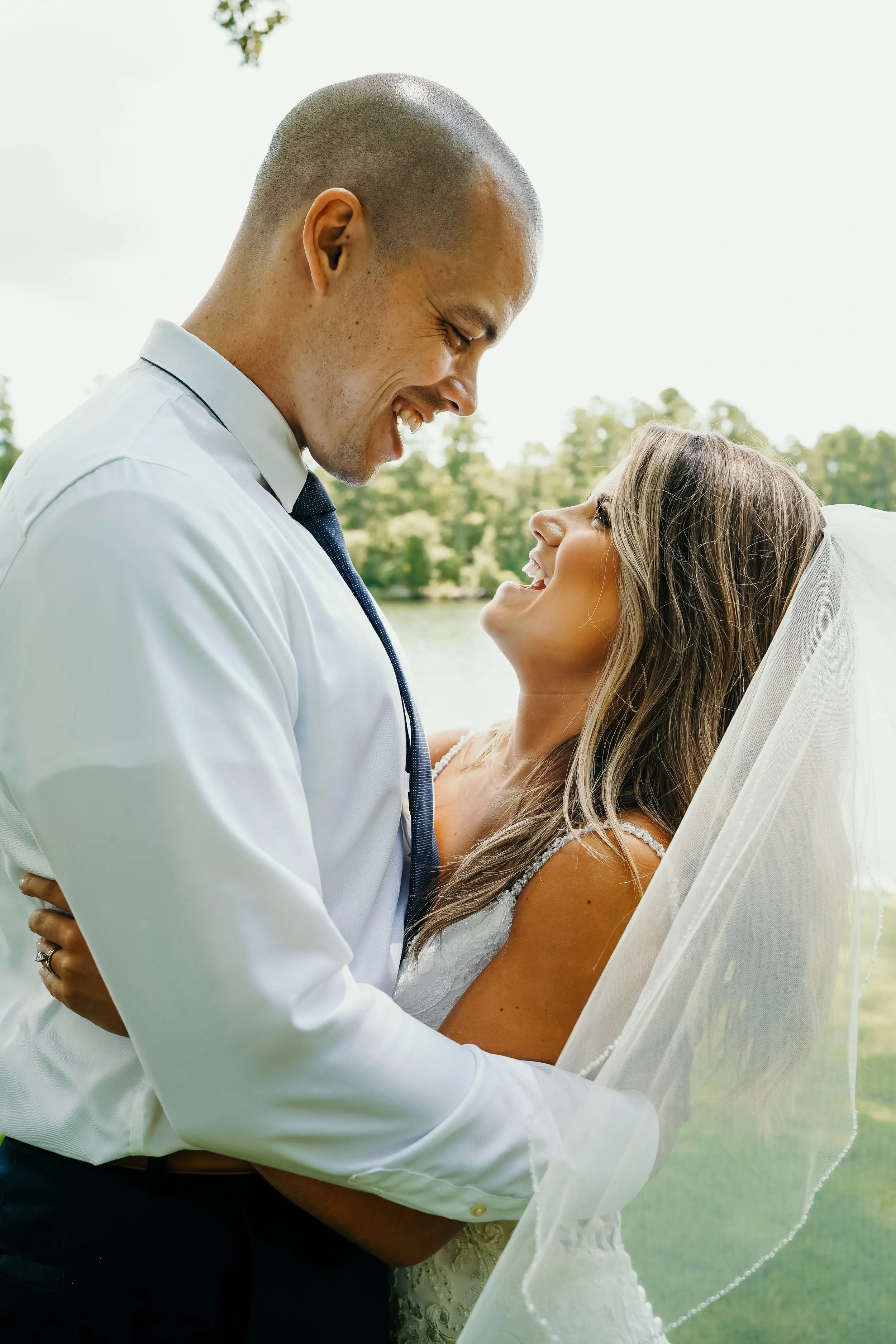 A couple on their wedding day, smiling and looking at each other outdoors near a lake with trees in the background.
