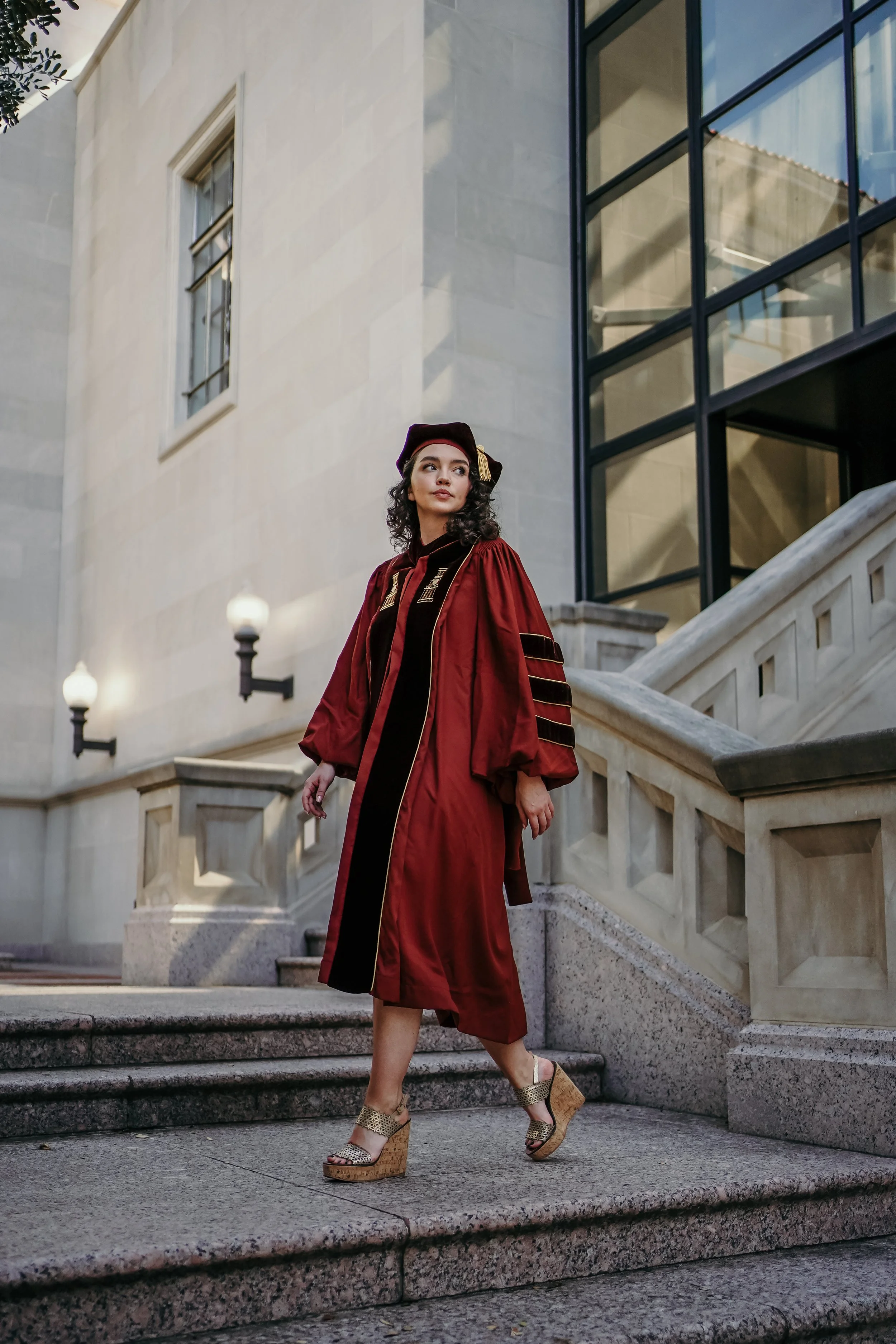 A young woman in a maroon graduation gown, matching cap, and gold and black heels walking down stone steps outside a building with large glass windows and stone walls.