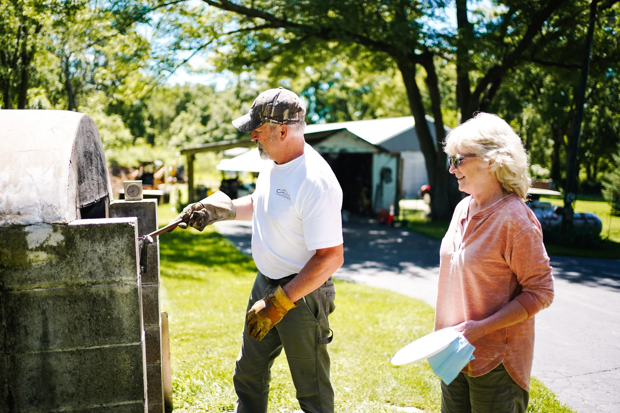 A man and woman outdoors on a sunny day are cleaning a large, weathered outdoor oven or grill. The man is wearing a baseball cap, white t-shirt, and gloves while scrubbing the oven. The woman, holding a plate and face mask, is smiling and observing h