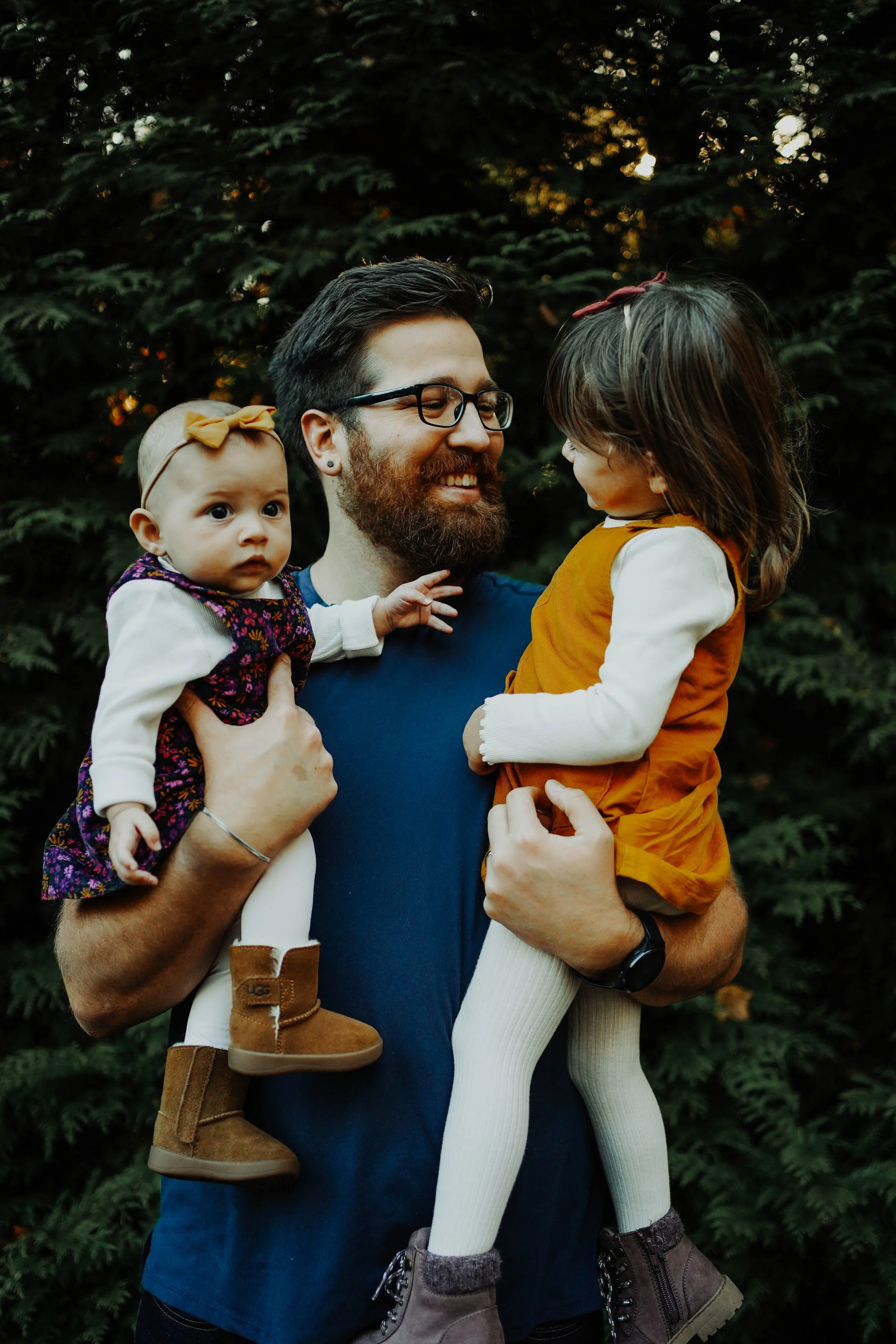 A bearded man wearing glasses holds two young girls, one in each arm, outdoors with green trees in the background. The girl on the left has a yellow bow in her hair and wears a colorful dress and tan boots. The girl on the right has a red bow in her 