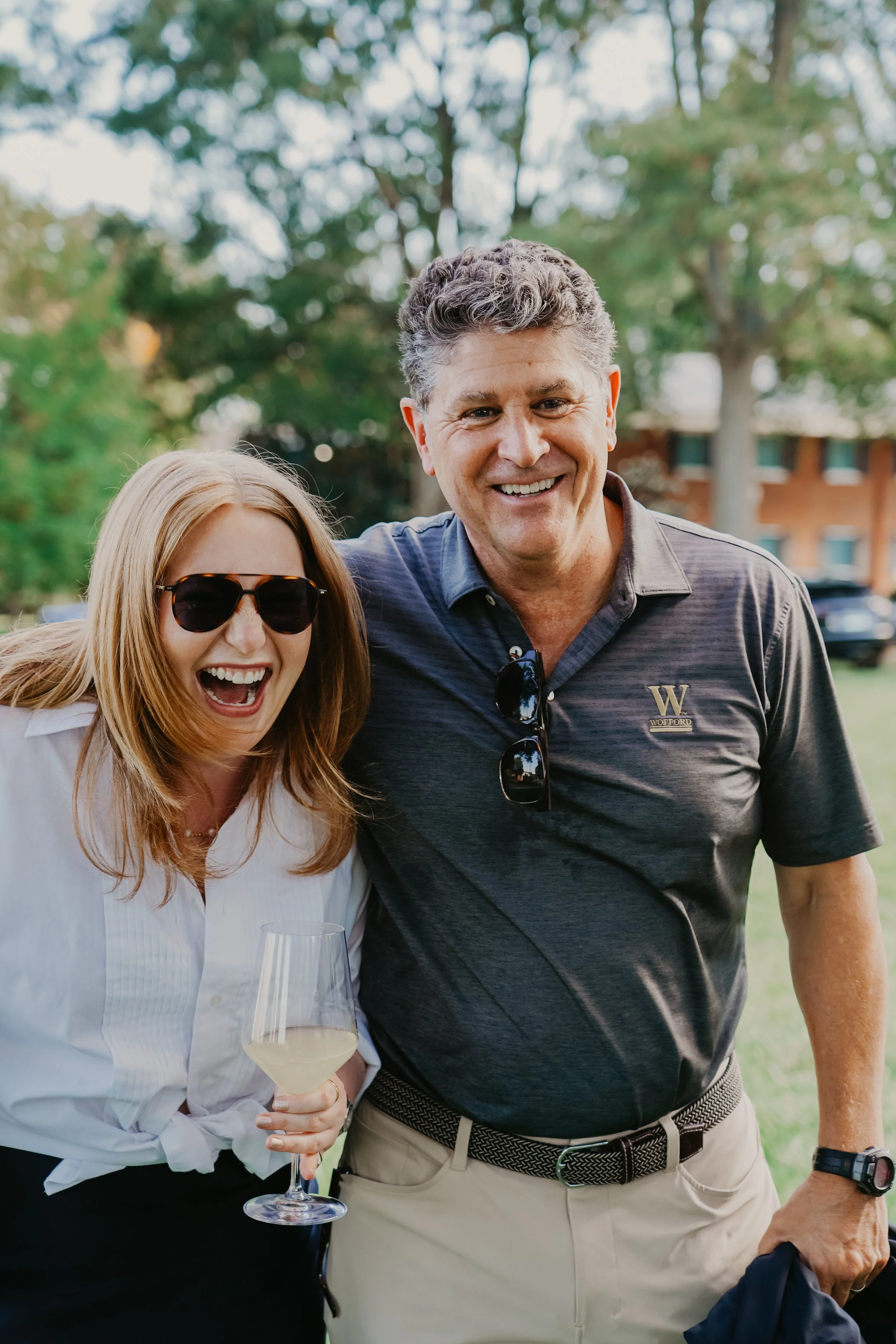 A man and woman smiling and posing outdoors, with the woman holding a glass of white wine.
