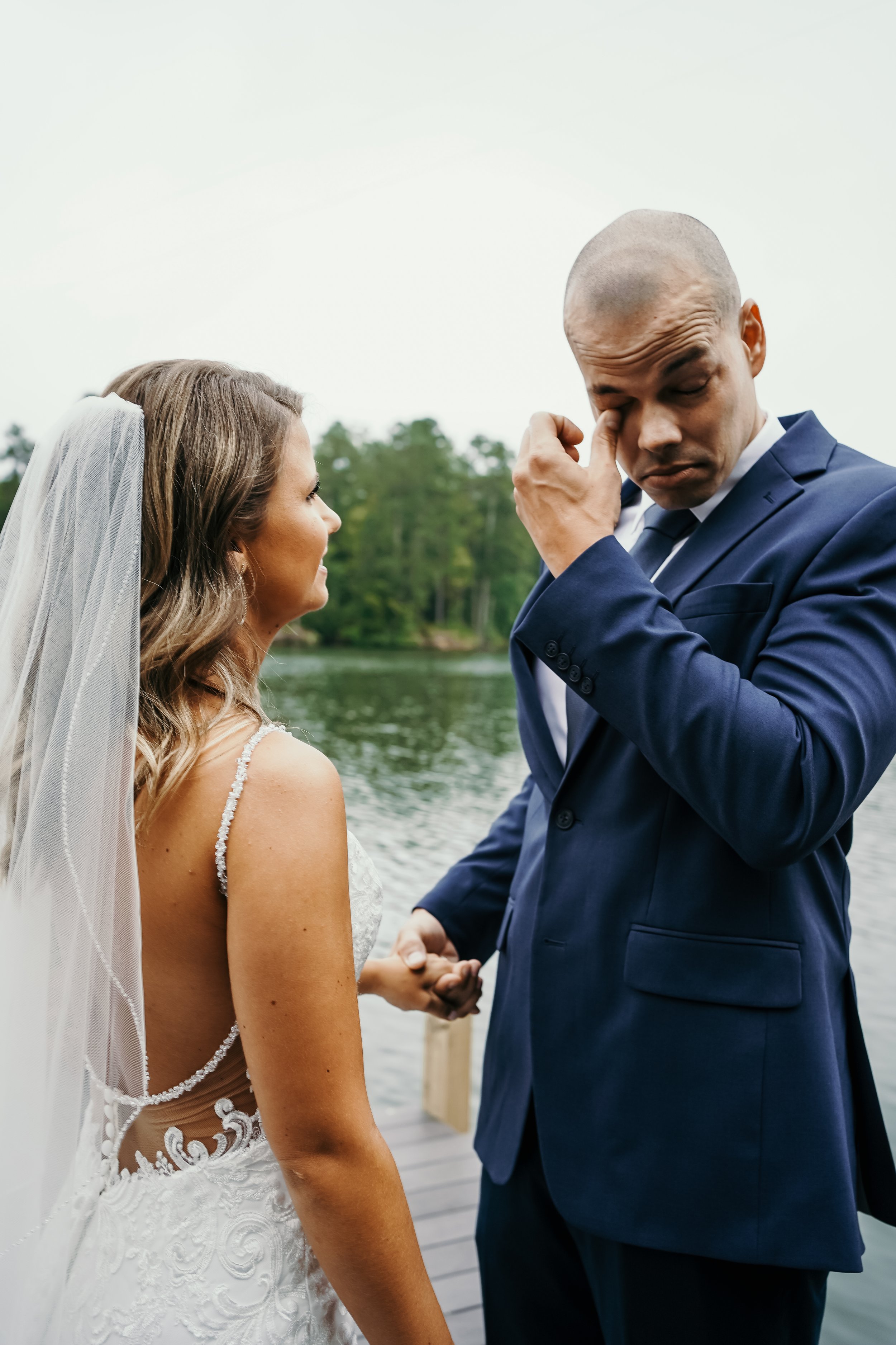 A bride and groom holding hands on a dock by a lake, with the groom wiping tears from his eyes.