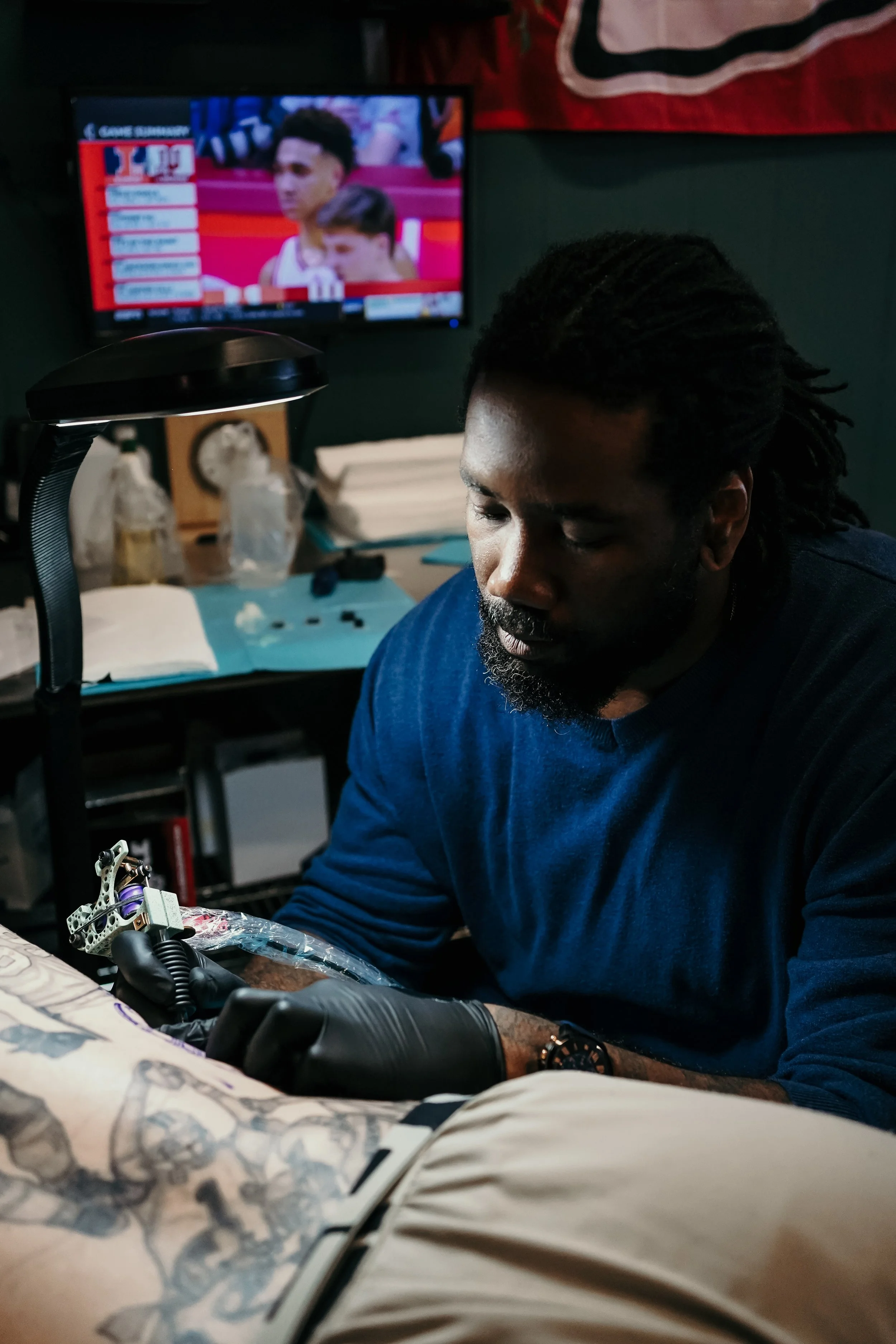 A man with dreadlocks and a beard working on a tattoo at a tattoo parlor. In the background, a television shows a sports broadcast, and there are various tattoo supplies on the table.
