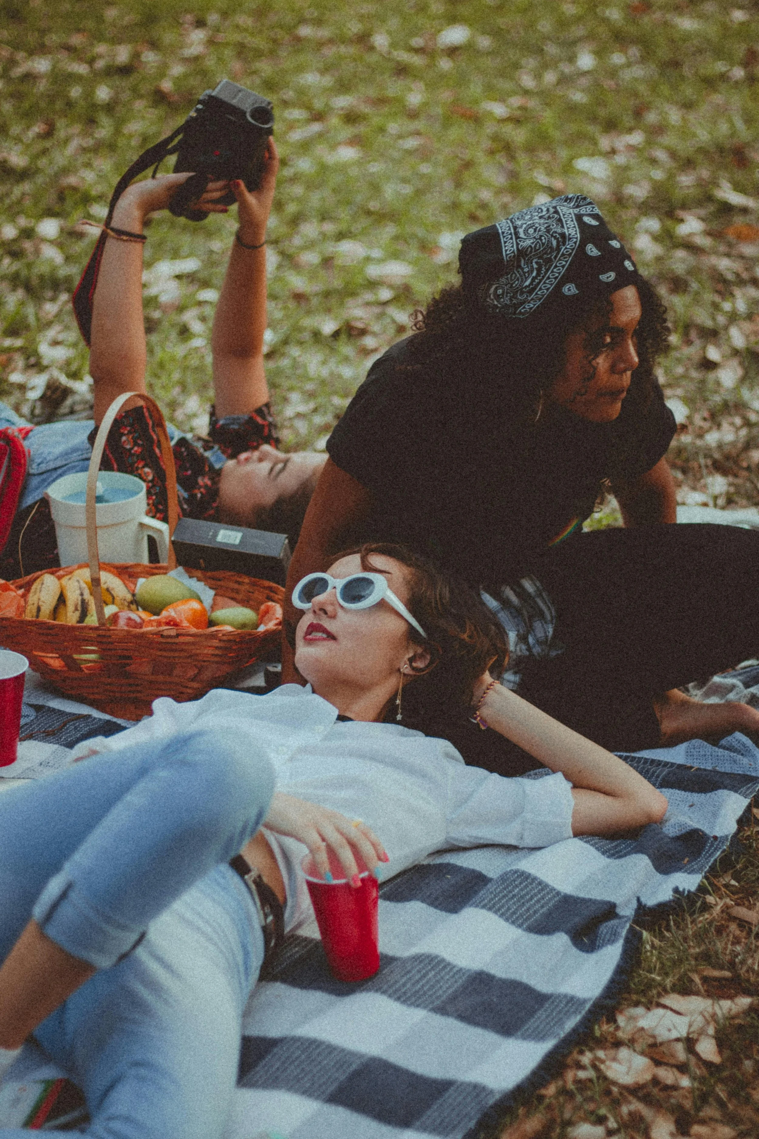 Three women having a picnic outdoors on a blanket; one woman is lying down with sunglasses, another woman is sitting and looking to the side, and the third woman is lying on her back holding a camera.