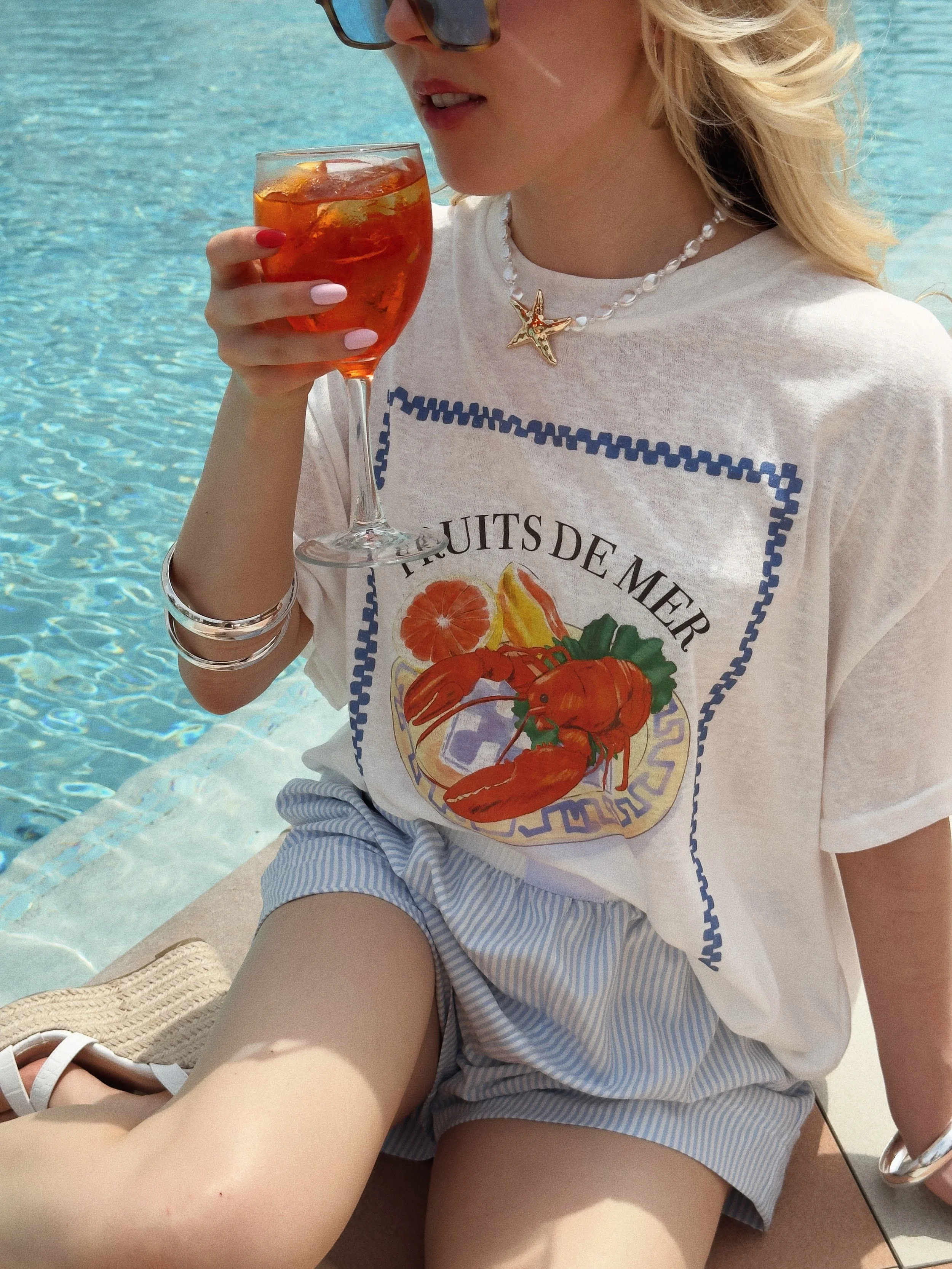 A woman sitting by a pool, holding a glass of iced tea with lemon slices, wearing sunglasses, a white t-shirt with a seafood print and French text, striped shorts, and accessories including a shell necklace and bangles.