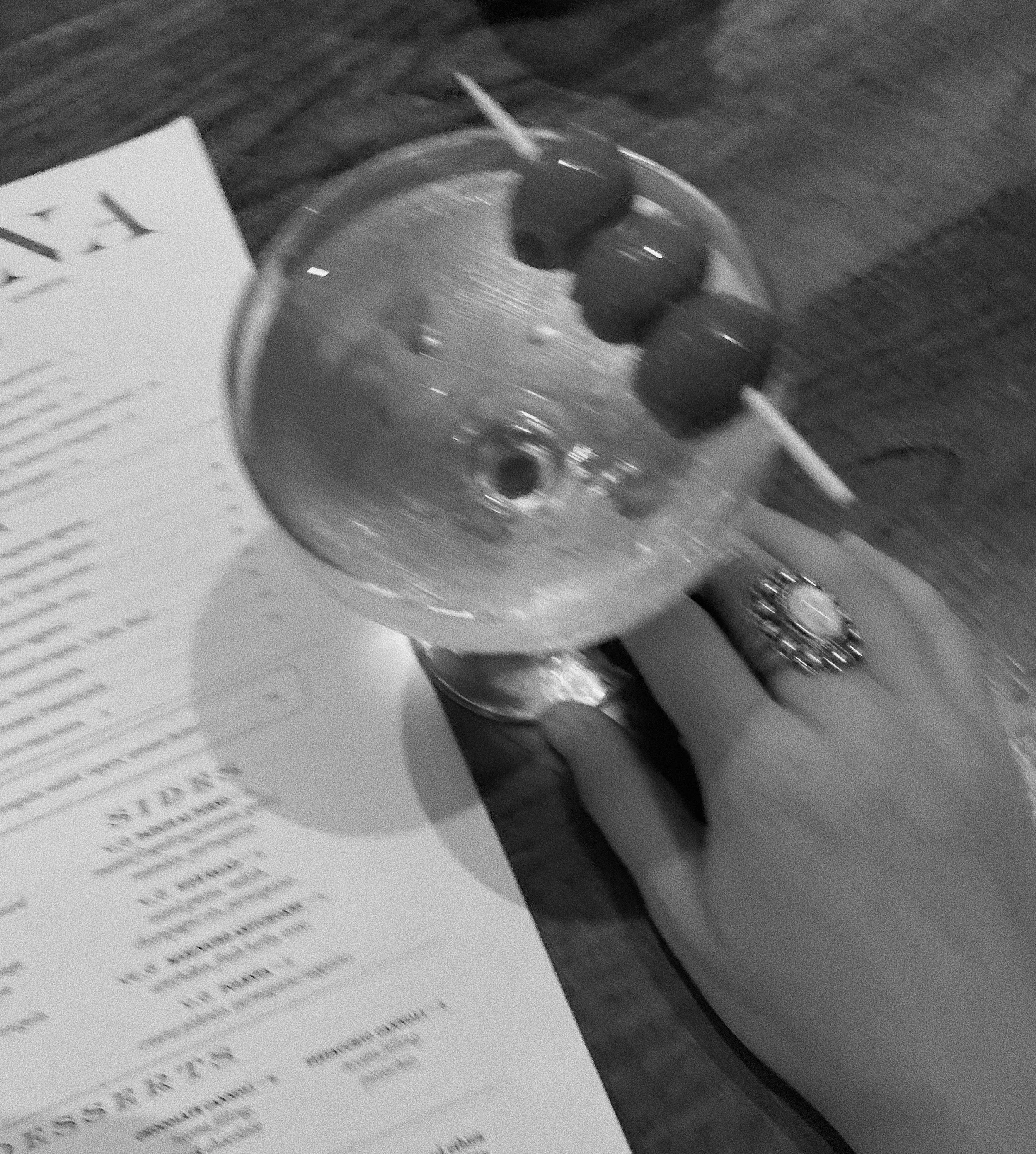 A hand with a ring on the ring finger holding a glass of water with three cherries on a toothpick, placed on a wooden table next to a printed menu.