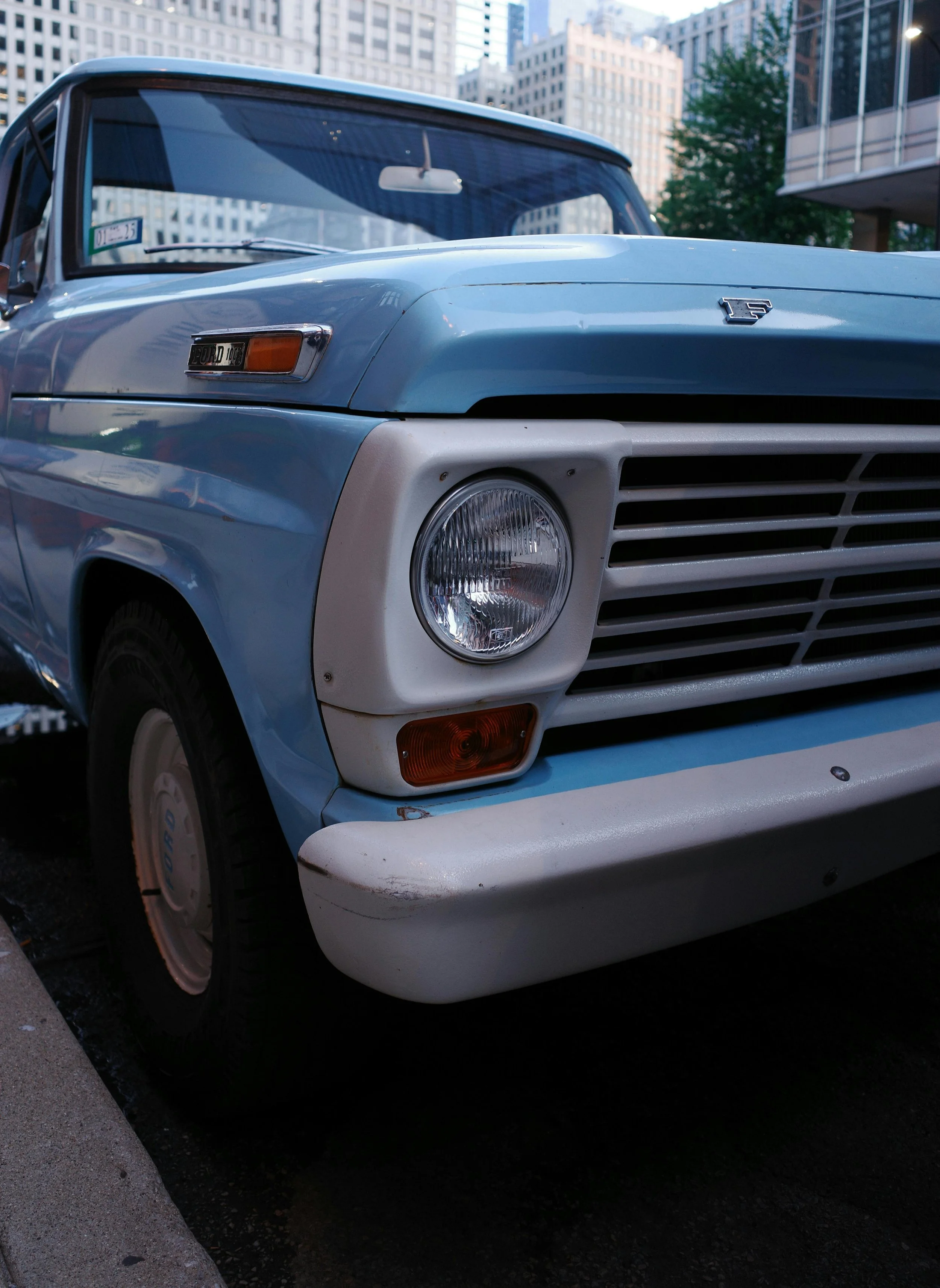 Close-up of a light blue vintage Ford truck, showing the front left side with round headlight, turn signal, and grille, parked in an urban area with tall buildings in the background.