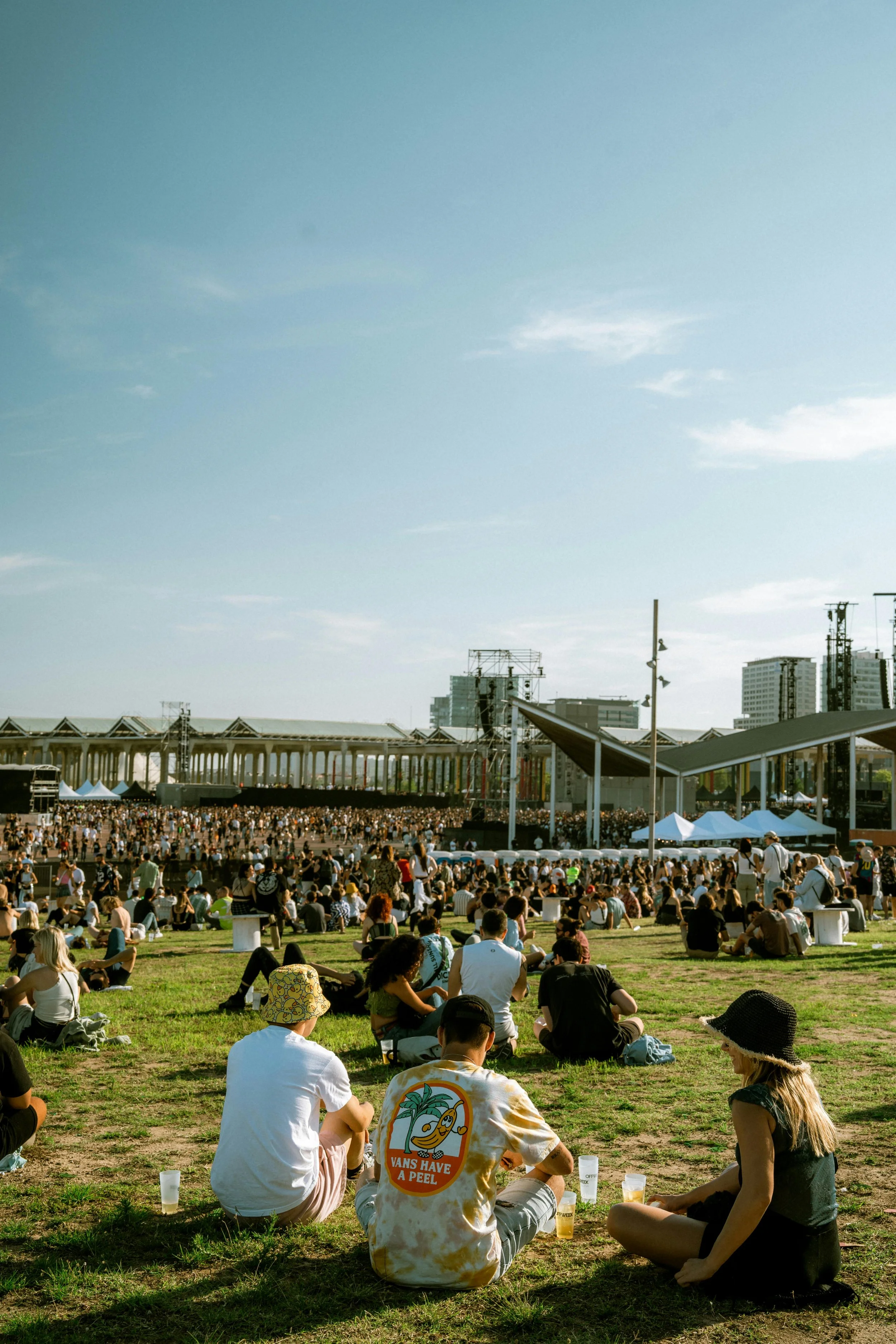 Crowd of people sitting on a grassy area watching a large outdoor concert or festival stage under a clear sky.