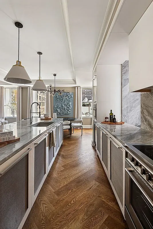 Modern kitchen with white cabinetry, gray marble countertops, and pendant lighting. A table with chairs is by a window in the background, with wall art and windows letting in natural light.