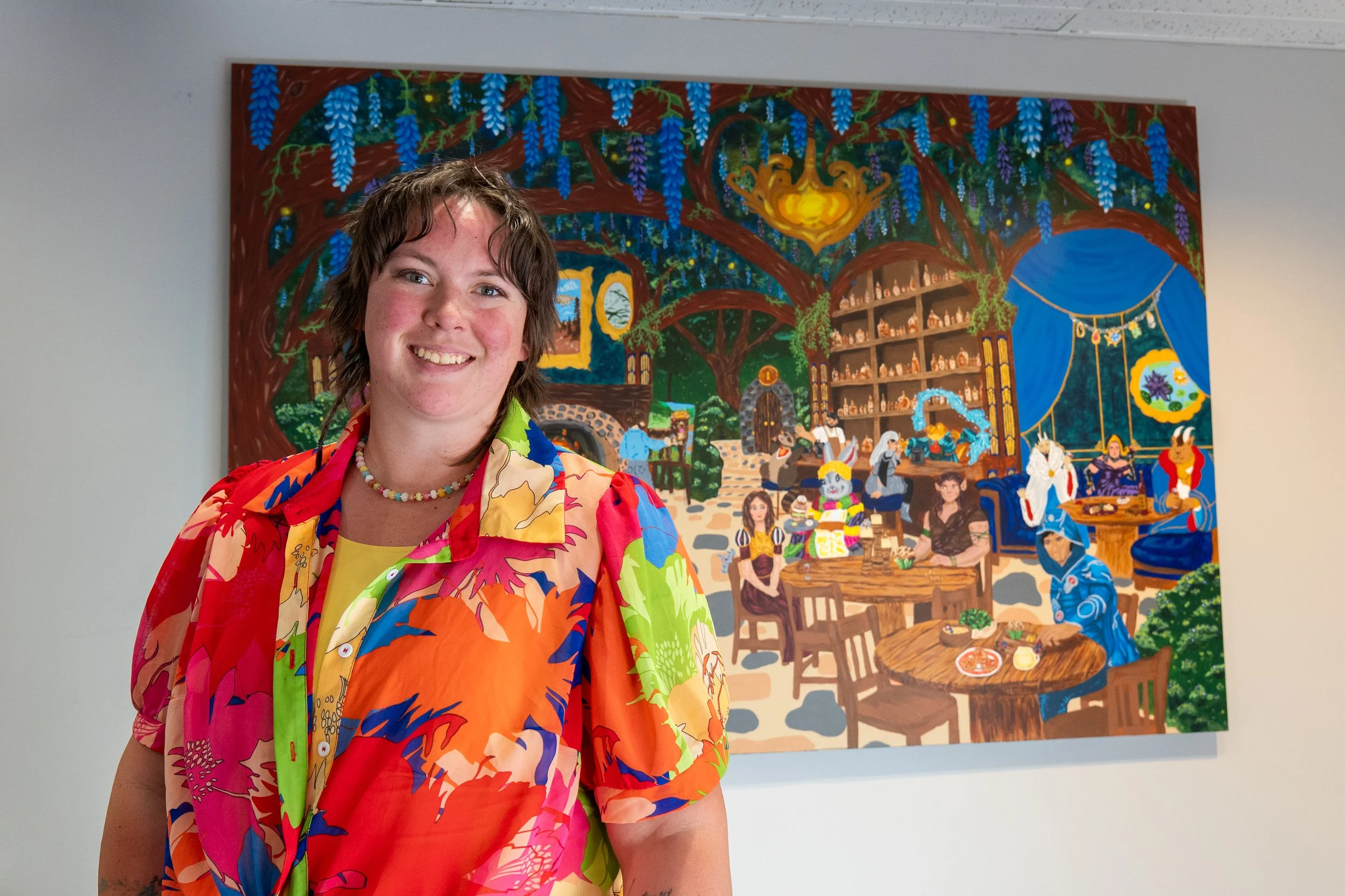 Artist Willow Jade standing and smiling in front of a colorful custom fantasy mural inside Syndicate Games in Utah, featuring whimsical woodland architecture, vibrant characters, tables, and magical details.