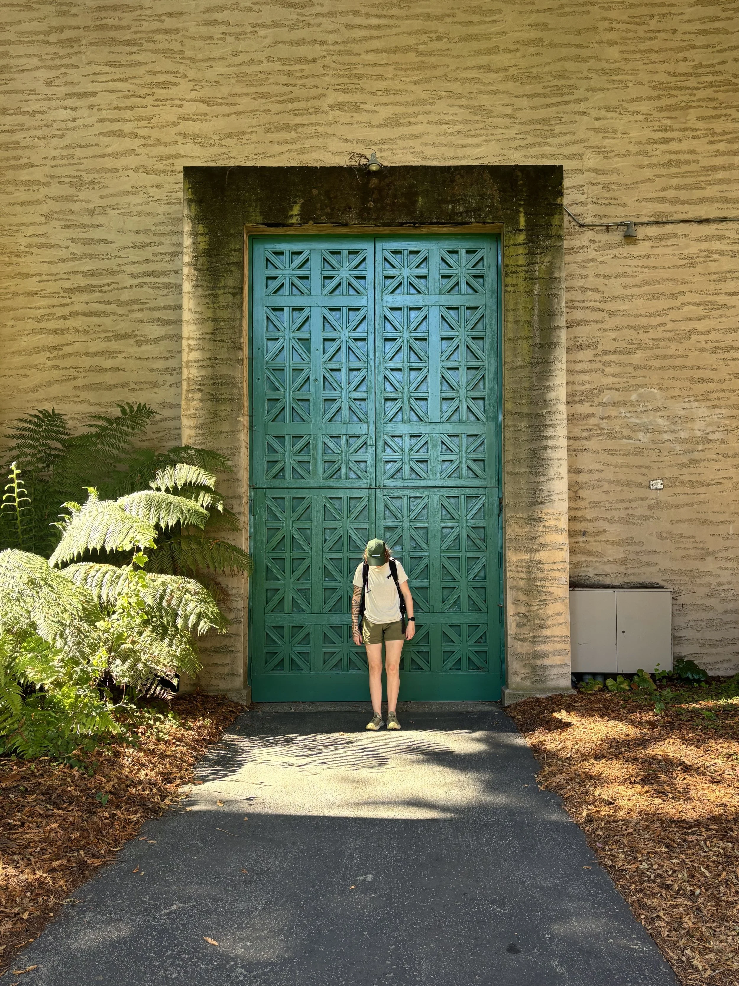 Wren Tilbrook Art, Ashbourne Derbyshire, Peak District. Person dressed in cream shirt, green shorts, and green cap standing in front of a dark green geometric patterned door built into a beige brick wall.