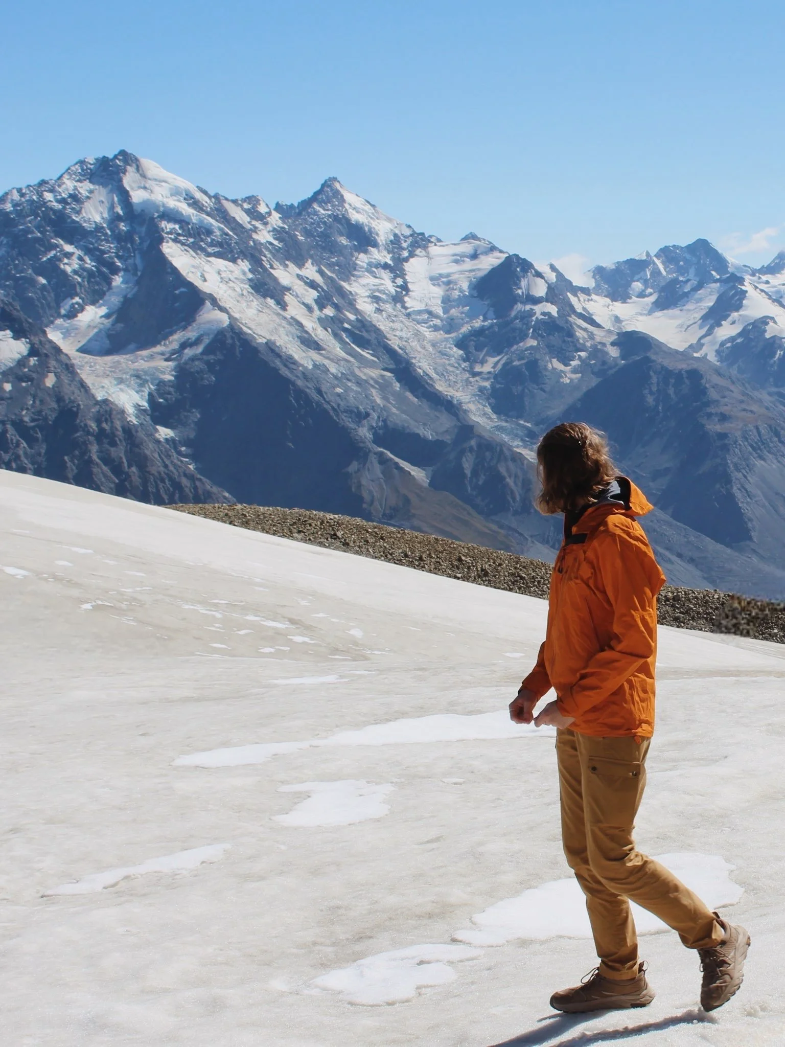 A person dressed in an orange jacket and tan pants walking on snow with snow-covered mountains in the background under a clear blue sky. Wren Tilbrook Art, Ashbourne Derbyshire, Peak District