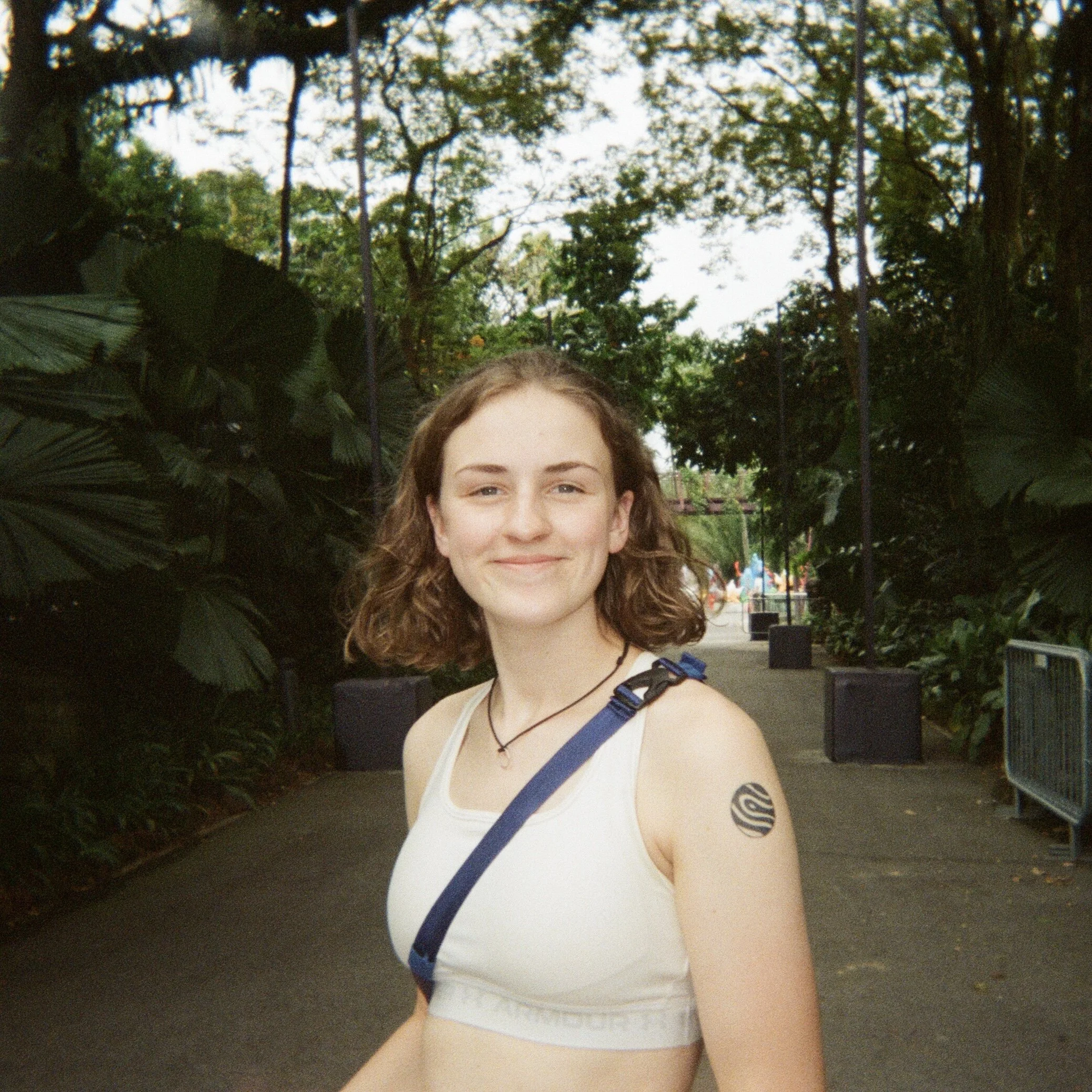 Young woman with curly brown hair smiling outdoors, wearing a white sports bra and a tattoo on her upper arm, in a park with trees and a pathway. Wren Tilbrook Art, Ashbourne Derbyshire, Peak District