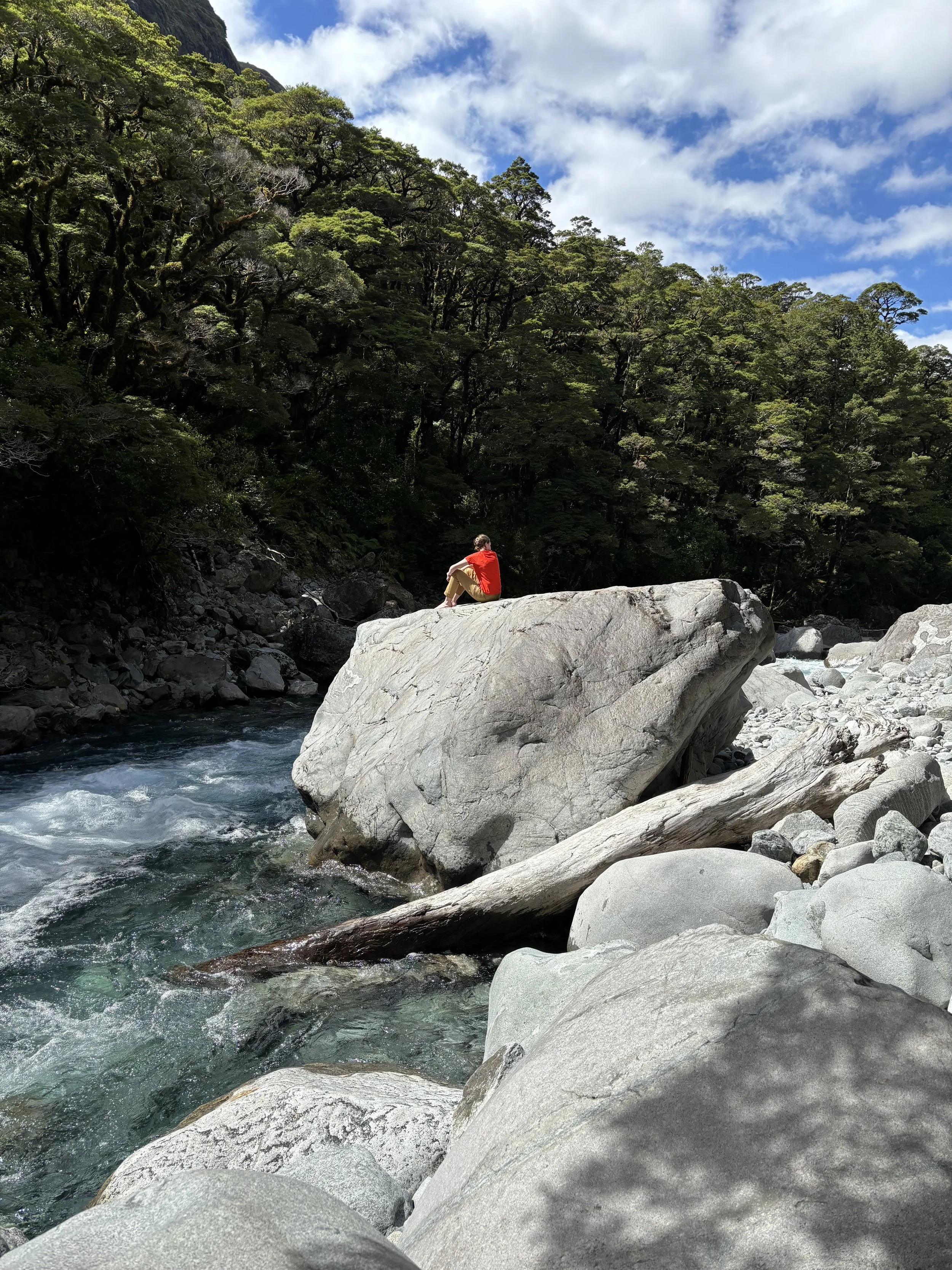 Person in a bright orange t-shirt sitting on a large gray rock by a flowing river, surrounded by trees and rocks under a blue sky with clouds. Wren Tilbrook Art, Ashbourne Derbyshire, Peak District