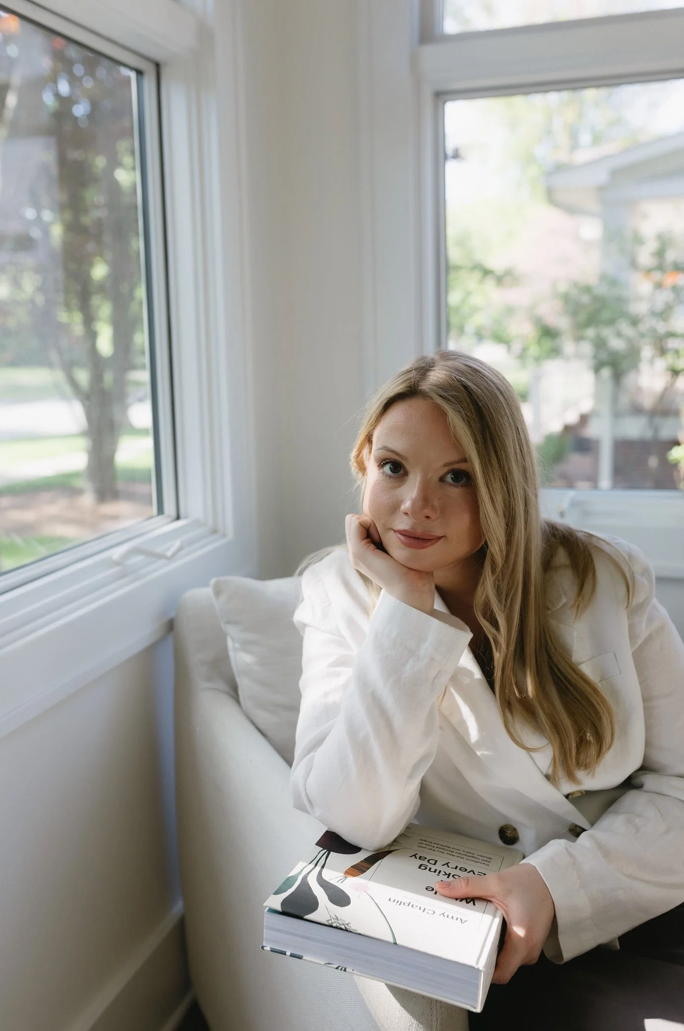 A woman with long blond hair sitting on a white couch, resting her head on her hand, holding a book titled 'Design Every Day' by Amy Chapman, next to a window with a view of trees and a house outside.