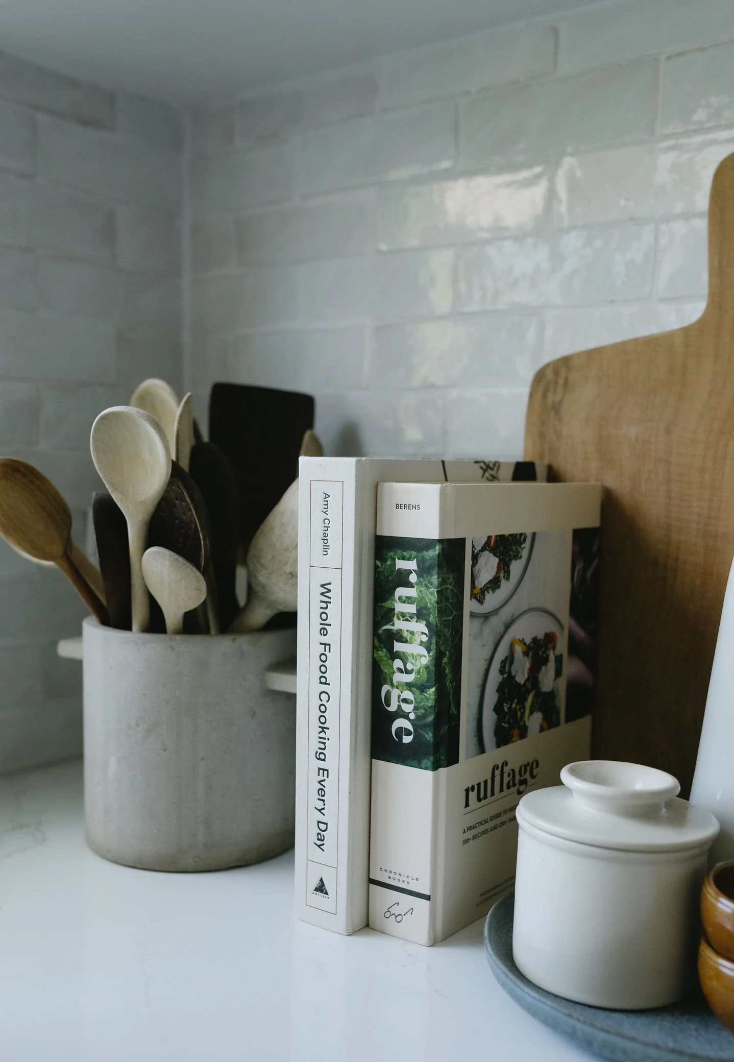 Cookbook titled 'Ruffage' and 'Whole Food Cooking Every Day' leaning against a brick wall, next to a white container filled with wooden and metal cooking utensils, and a small white lidded jar on a gray tray.