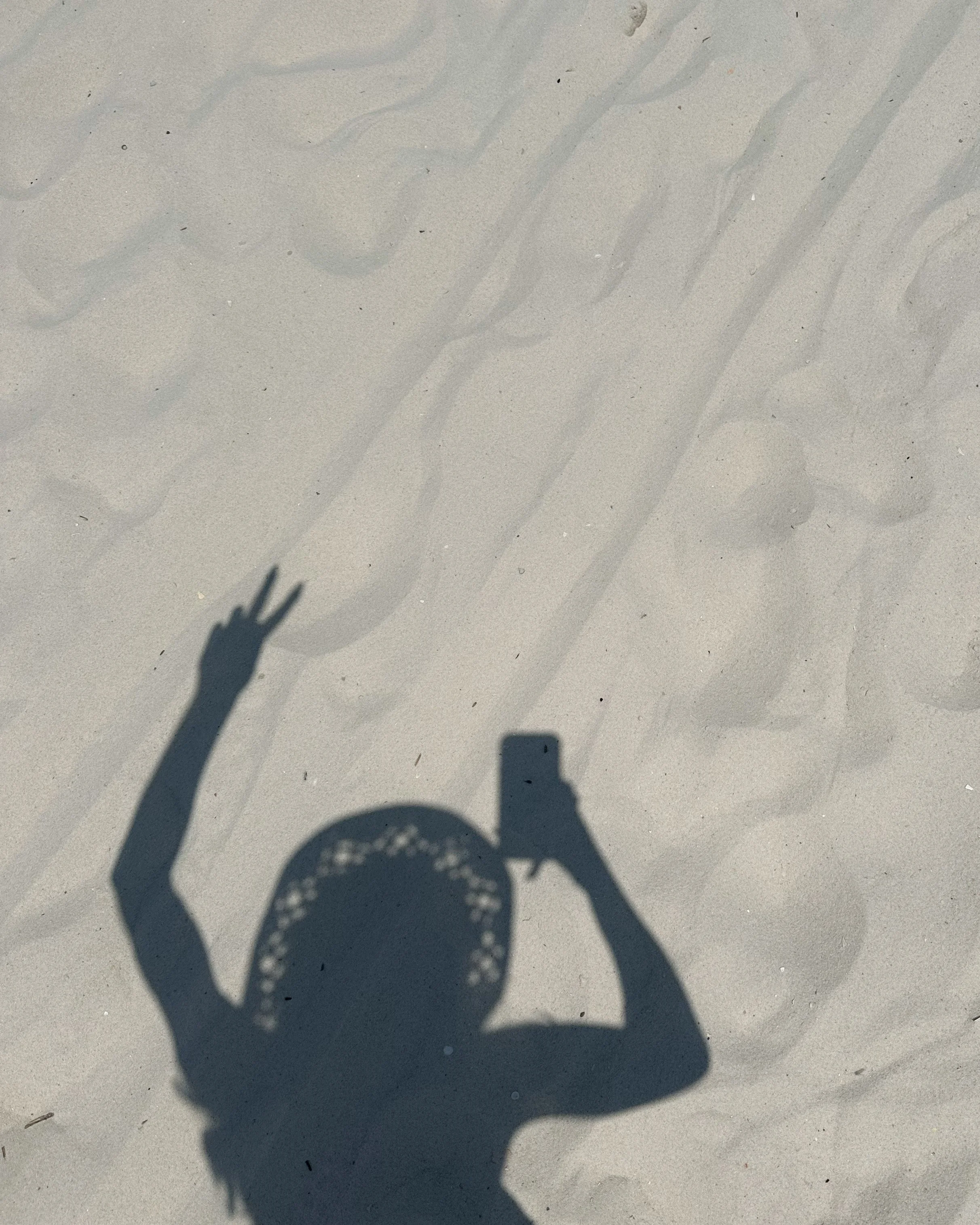 Shadow of a person taking a photo with a smartphone on the sand, with one hand raised and making a peace sign, with faint footprints in the sand.