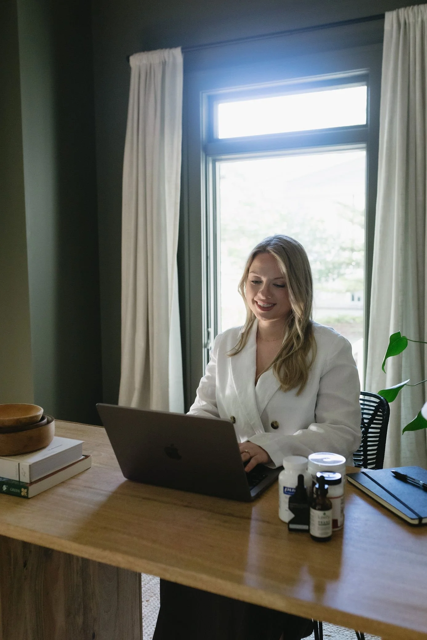 A woman sits at a wooden desk using a laptop, with a window behind her allowing natural light into the room. The desk has books, medication bottles, and a notebook on it.