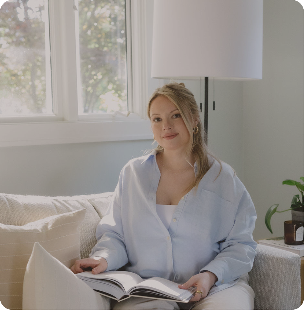 A naturopathic doctor, woman with blonde hair and hoop earrings sitting on a sofa holding a book in a sunlit living room with a window and a potted plant.