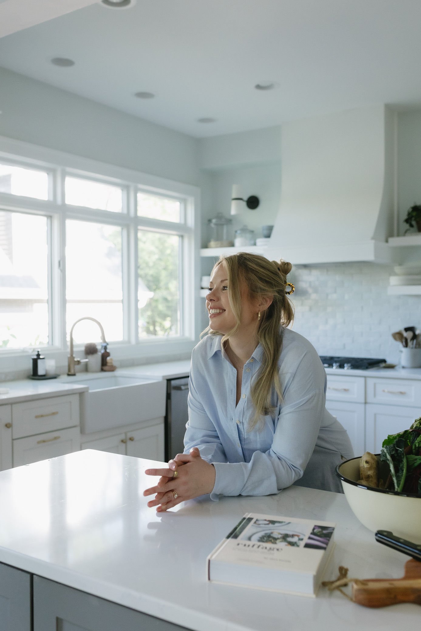 A woman with long blonde hair smiling and sitting at a white kitchen island in a bright, modern kitchen with large windows.