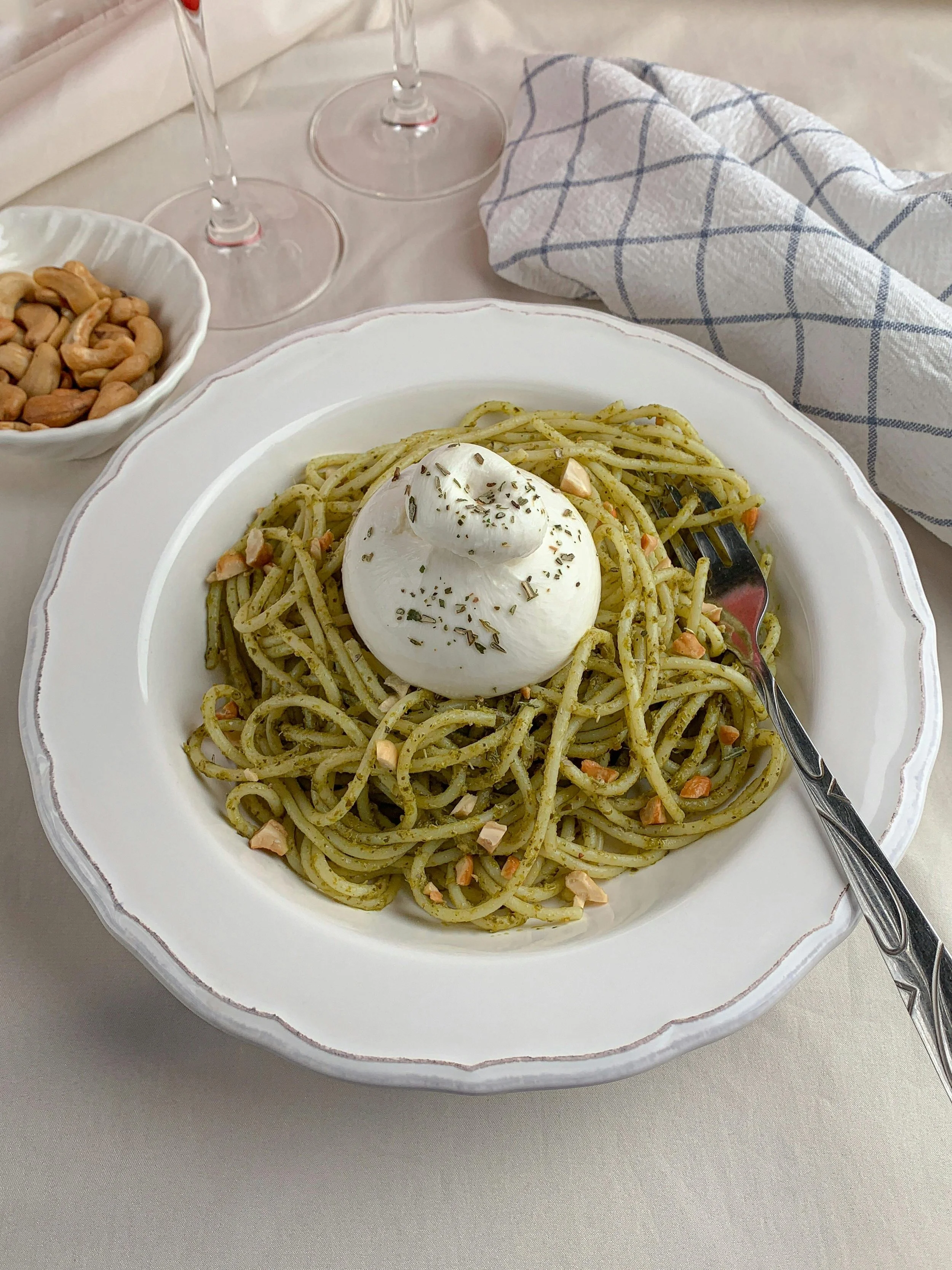 A plate of spaghetti with pesto sauce topped with a dollop of burrata cheese, garnished with dried herbs, on a white plate with a silver fork. In the background, there are two empty wine glasses, a small bowl of cashews, and a blue and white checkered kitchen towel.