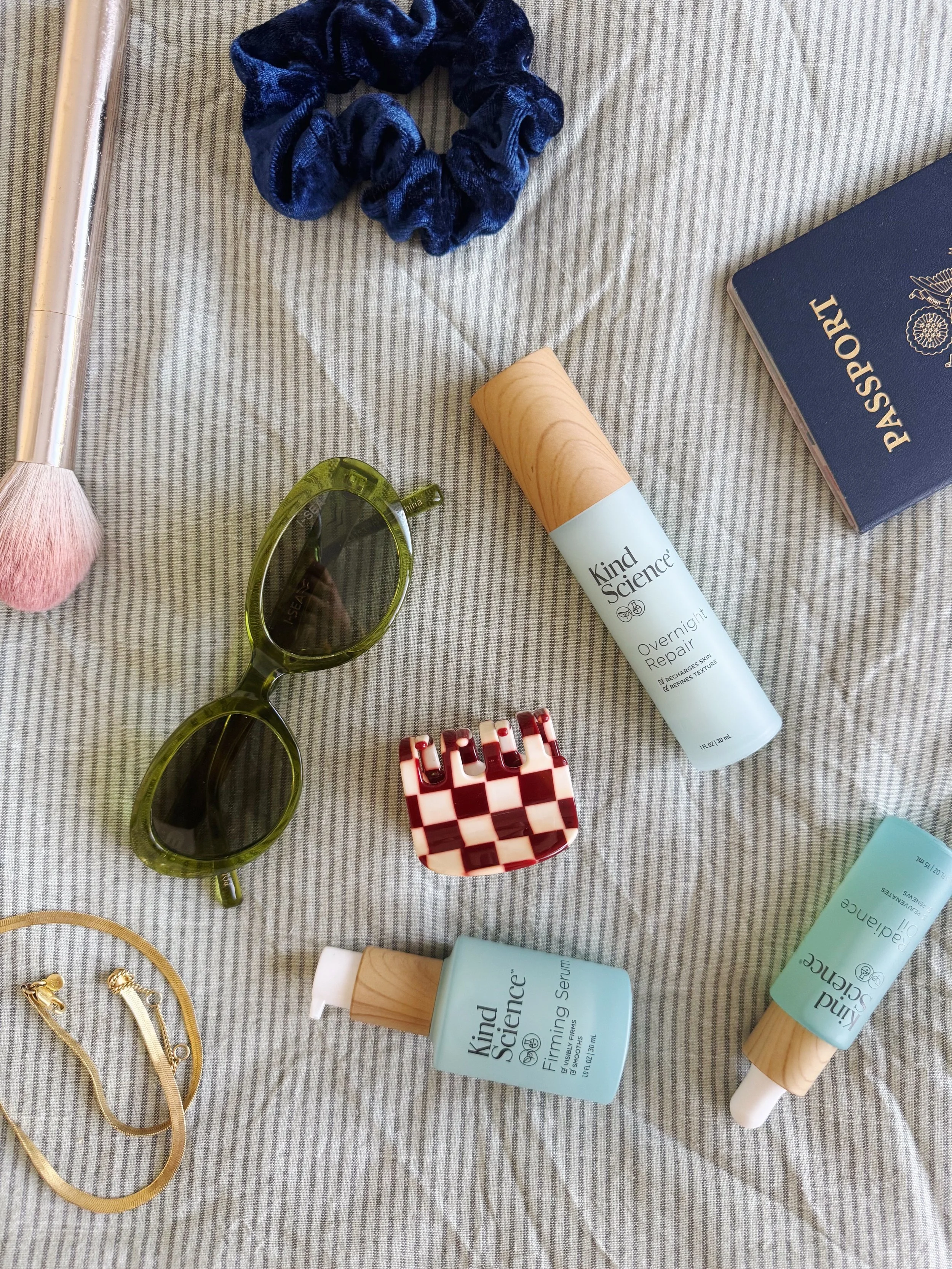 Travel essentials on a gray striped fabric including a navy blue scrunchie, green sunglasses, a red and white checkered hair clip, a gold necklace, a blue passport, and skincare products from Kind Science such as Firming Serum and Overnight Repair.