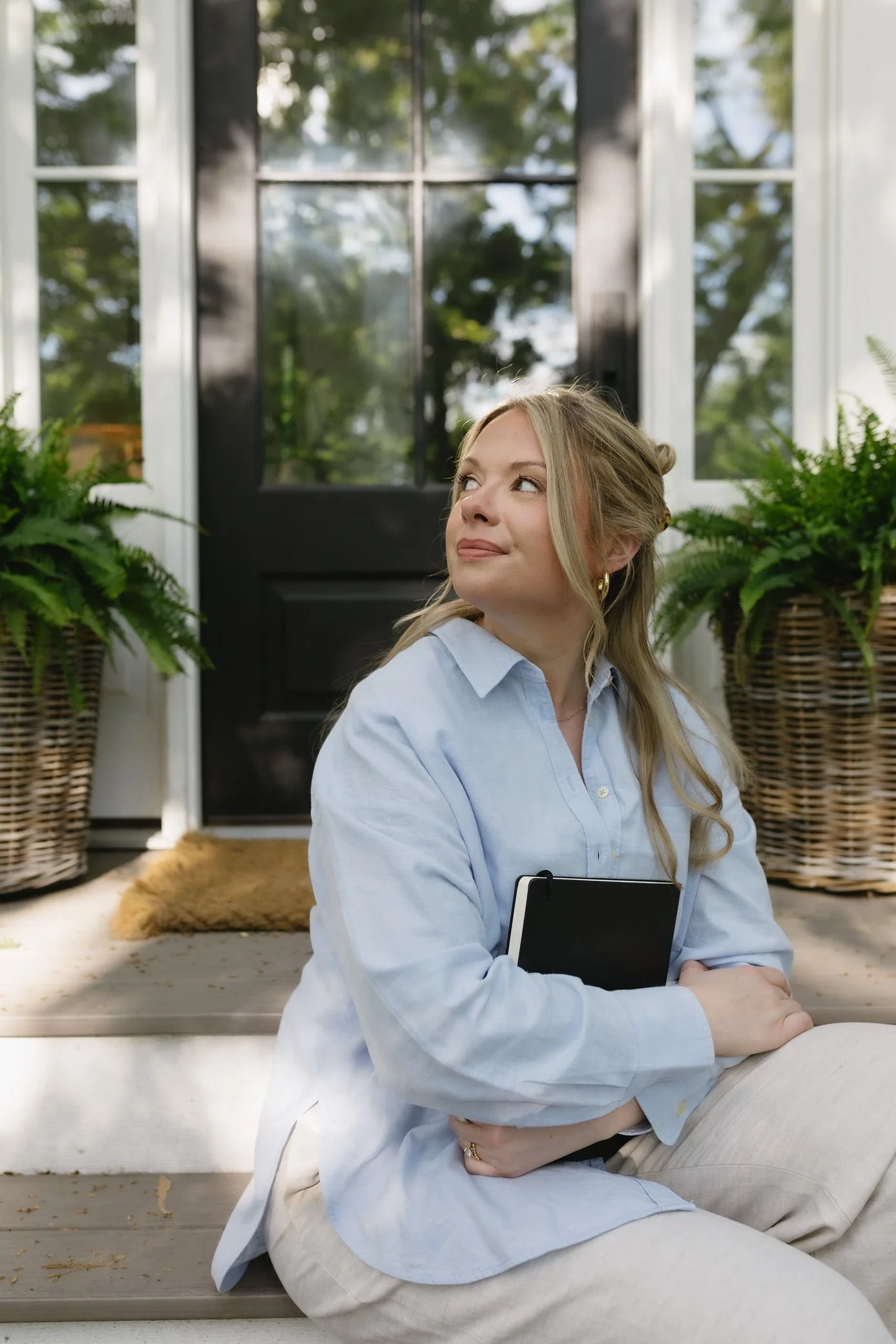 A woman sitting on the porch, holding a tablet, looking thoughtfully to the side with a smile, surrounded by potted ferns and a black door in the background.