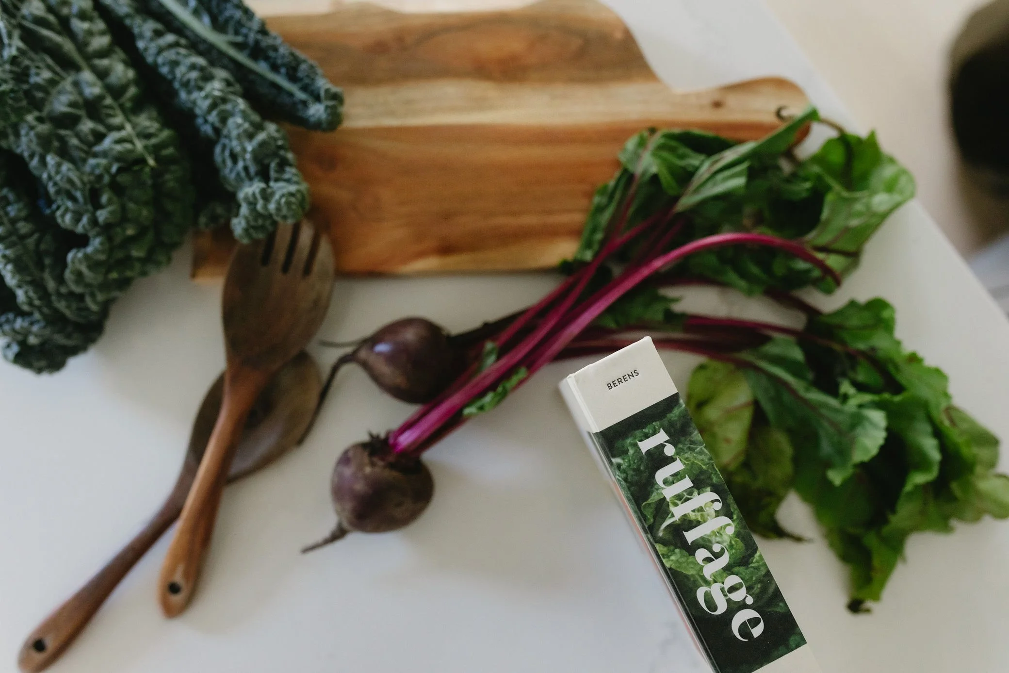 Fresh kale, beets with greens, and a package of arugula on a kitchen counter, with a wooden cutting board and two wooden utensils.