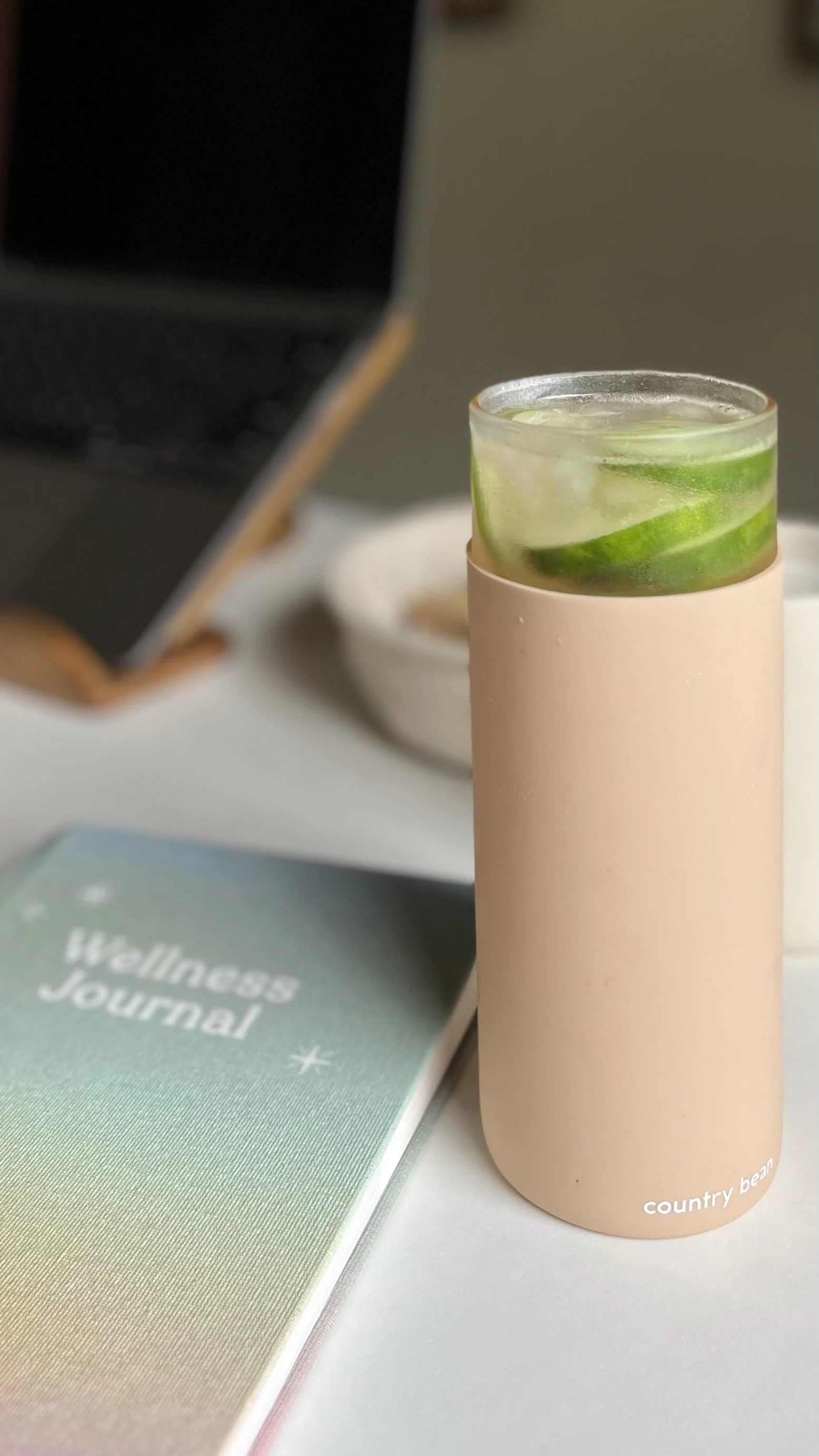 A tall beige tumbler with a glass of lime and ice inside, placed on a white surface with a Wellness Journal nearby.