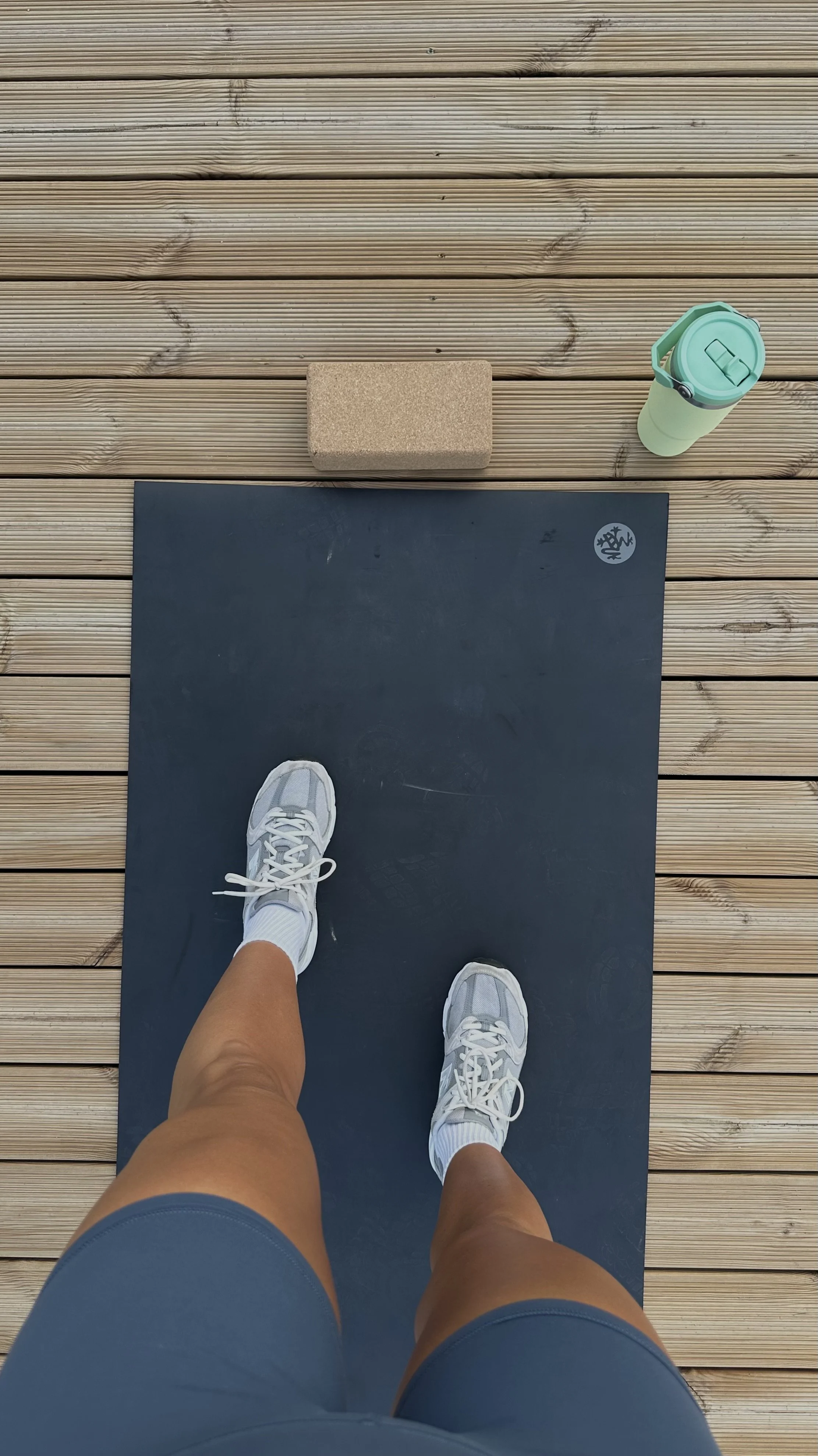 Overhead view of a person standing on a black yoga mat outdoors on wooden decking, wearing white sneakers and shorts, with a foam roller, a cork block, and a reusable travel mug nearby.