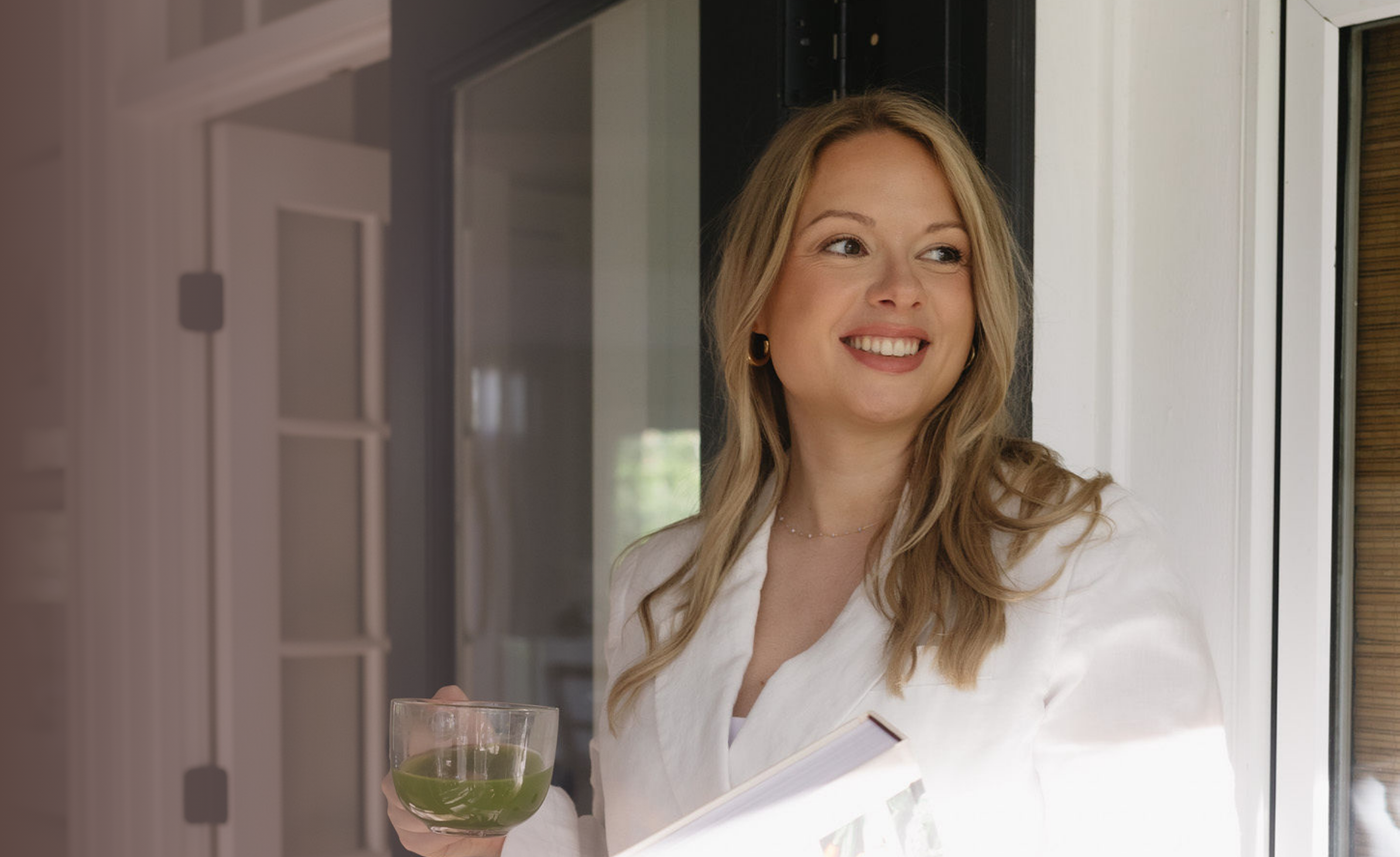 A woman with blonde hair, wearing a white blazer, holding a glass of green liquid, smiling and looking outside through a window.