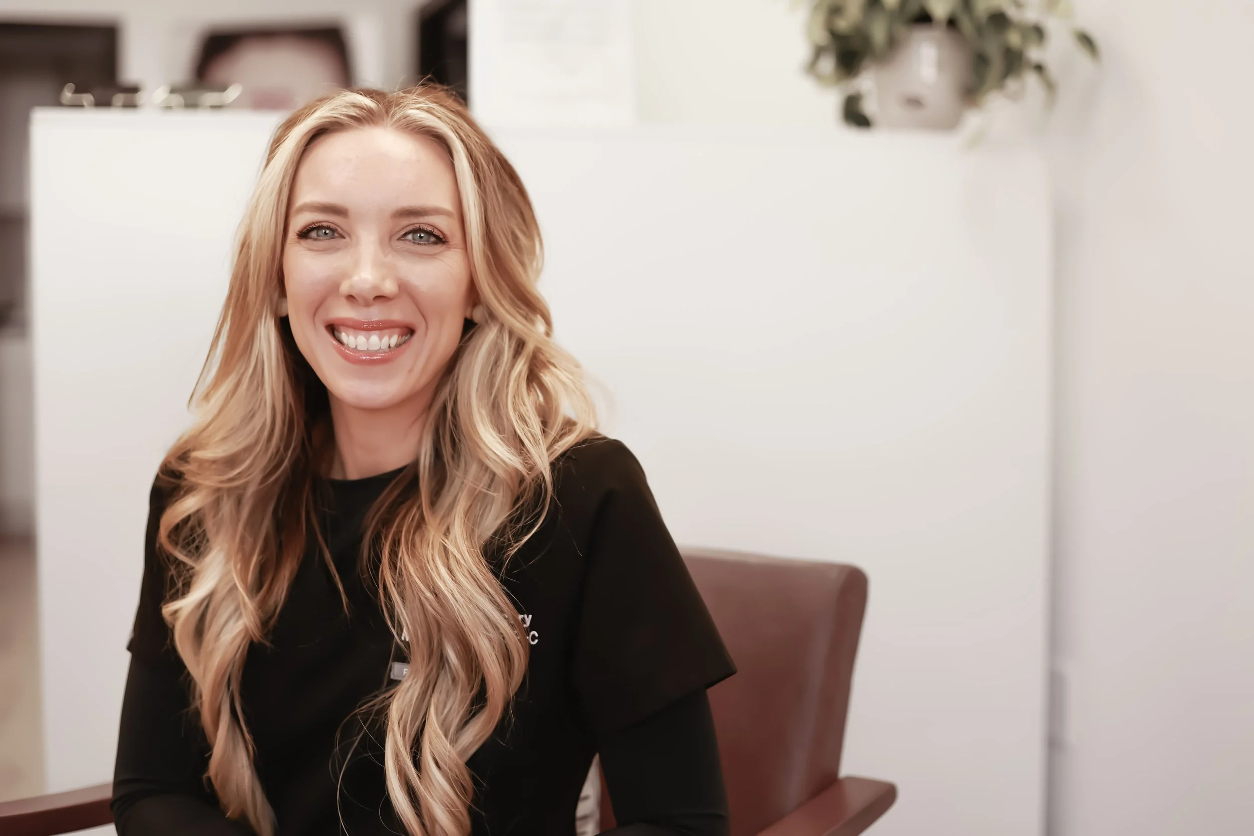 A smiling woman with long blonde hair sitting in a chair in a medical or office setting.