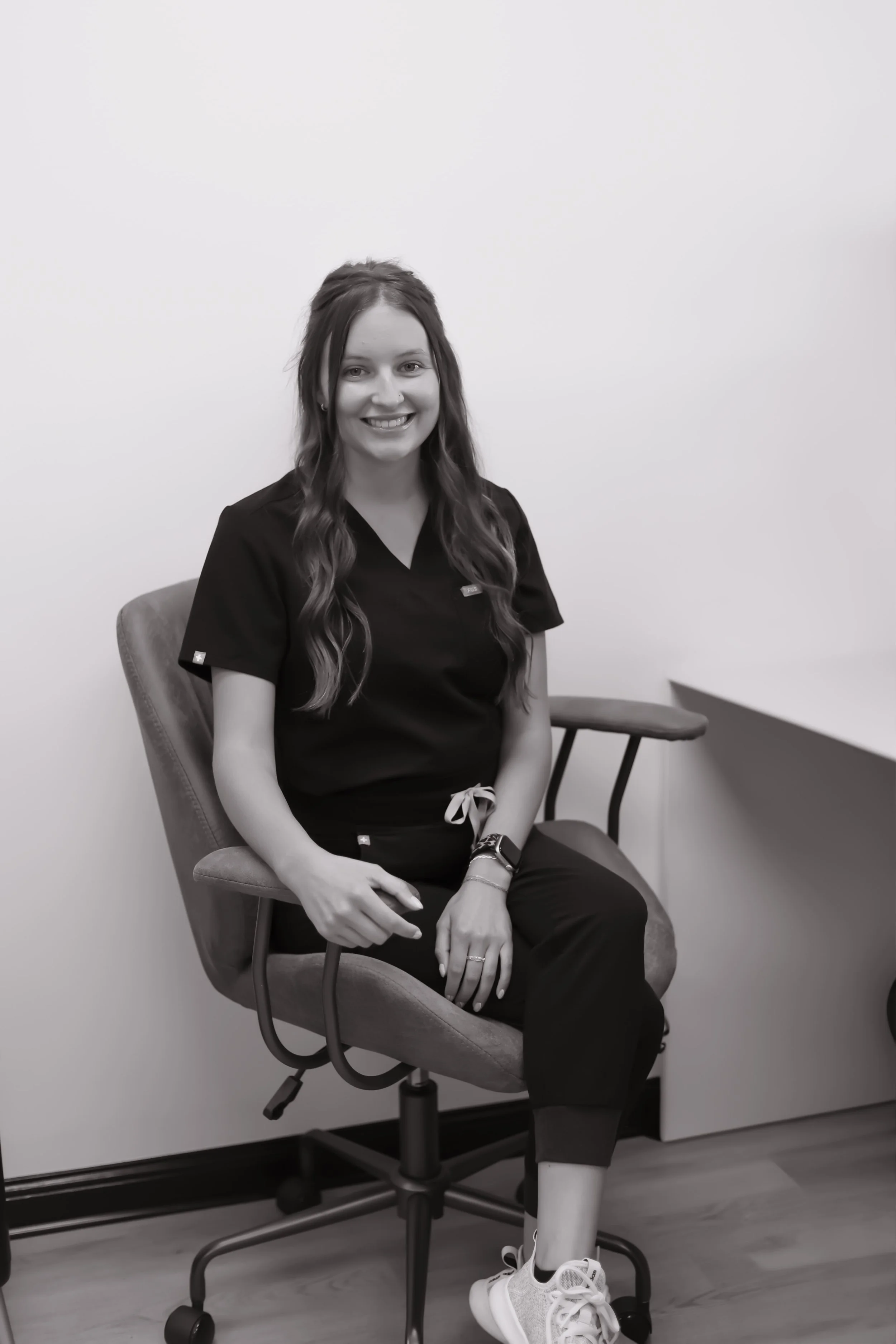 A young woman smiling, wearing medical scrubs, sitting on an office chair in a plain room.