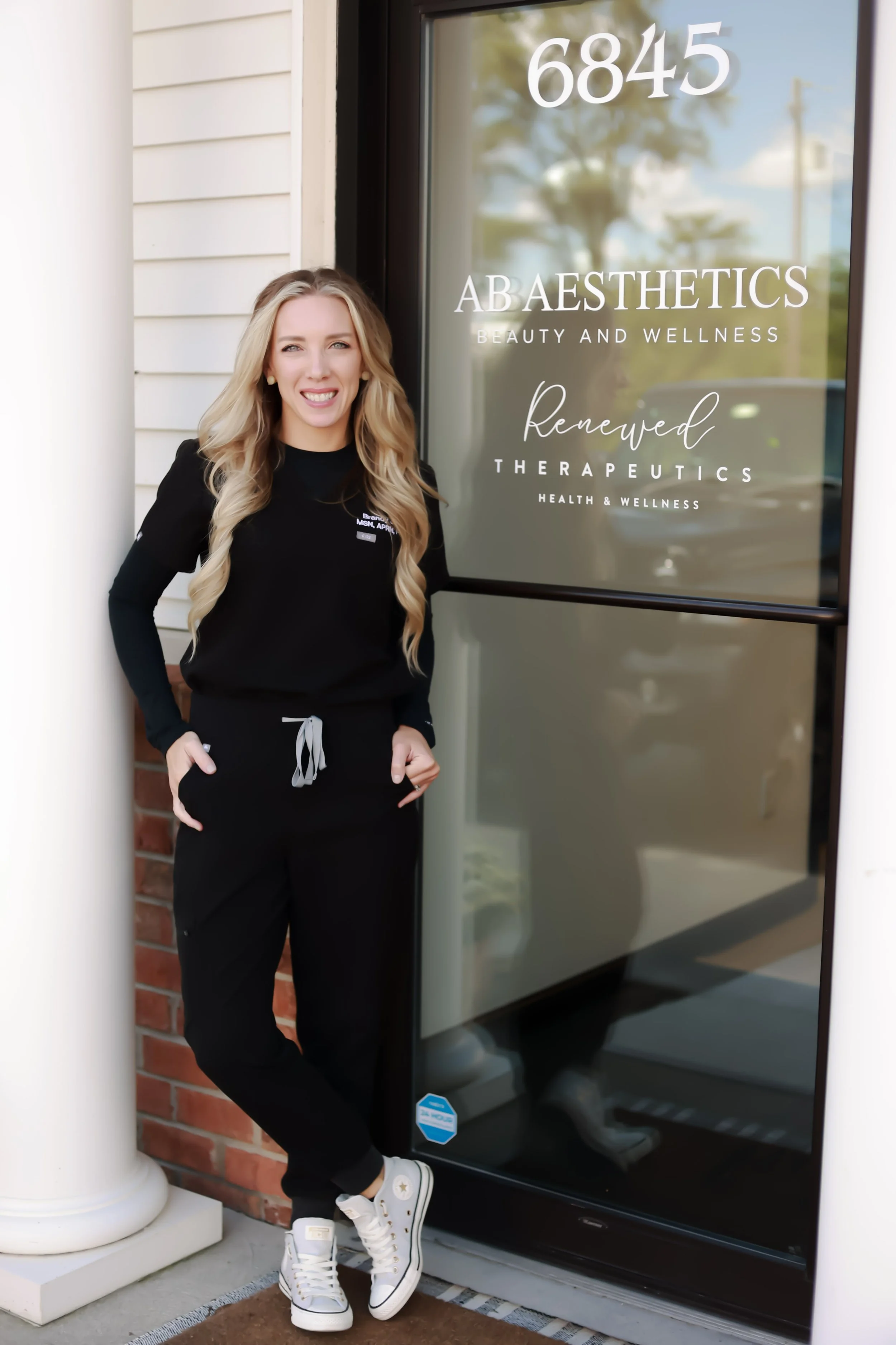 A woman with long blonde hair, wearing a black athletic outfit and white sneakers, standing outside a beauty and wellness clinic named A B Aesthetics, smiling at the camera.
