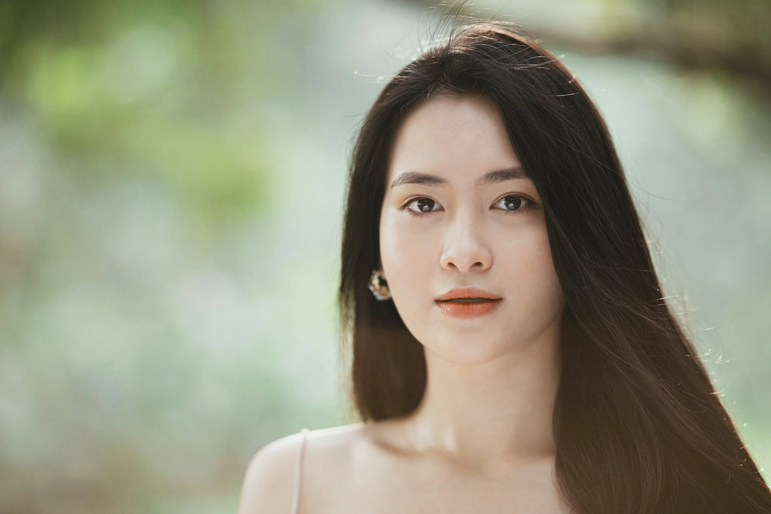 A portrait of a young woman with long dark hair, wearing a white top and earrings, outdoors with blurred greenery in the background.