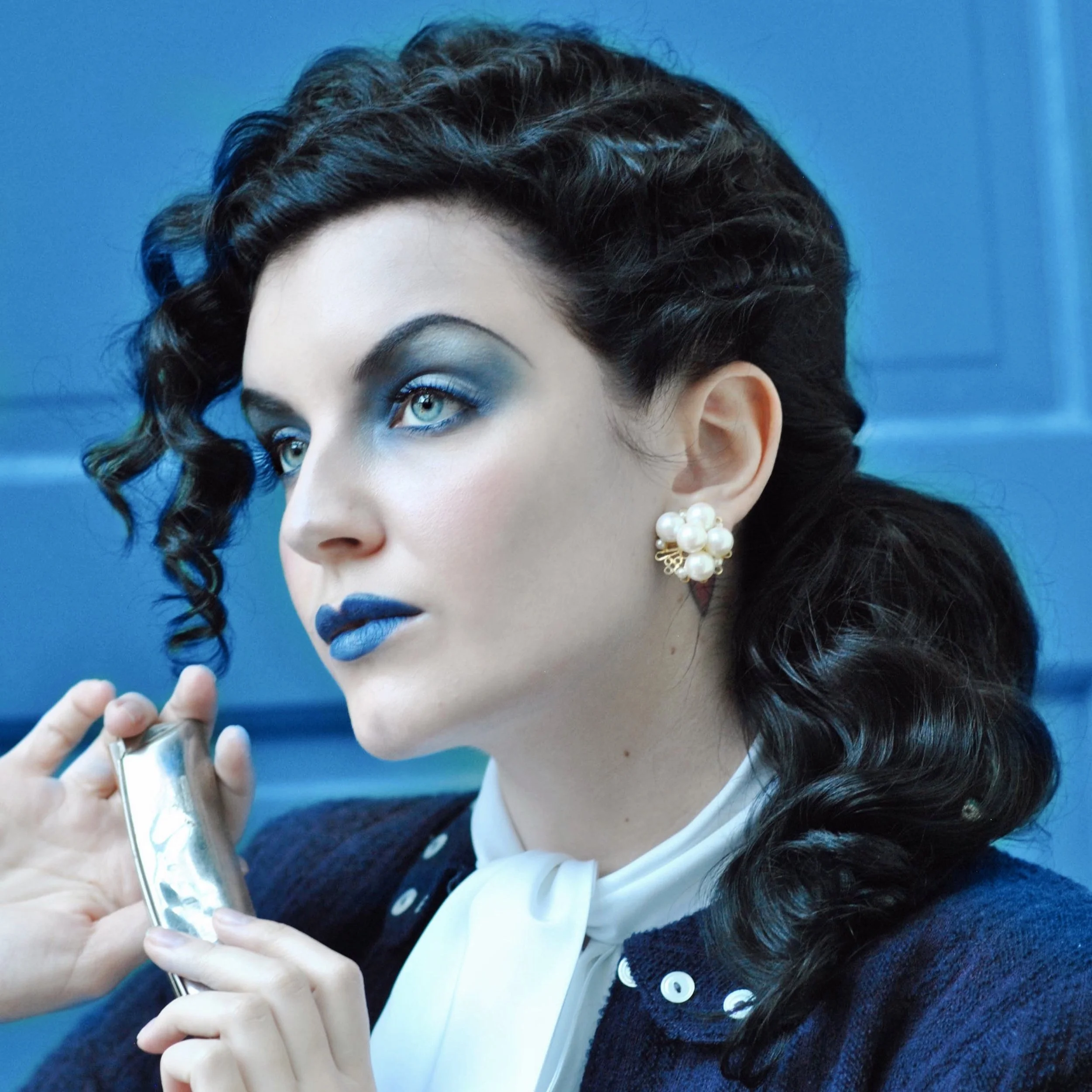 A woman with dark, curly hair and blue eye makeup holding a shiny rectangular compact mirror, wearing pearl earrings, a white neck scarf, and a dark sweater.