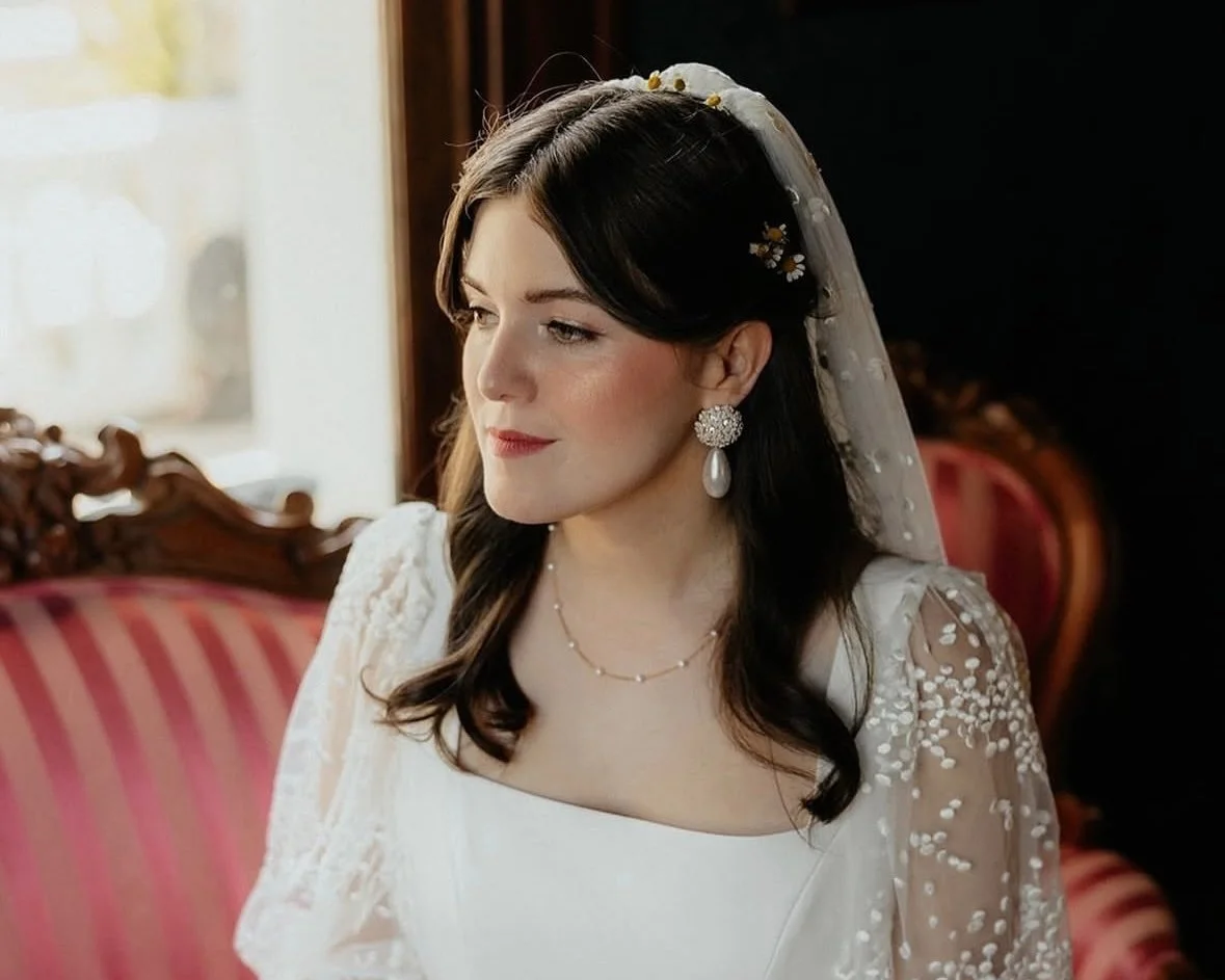 A woman with dark hair wearing a wedding dress, veil, and pearl earrings, sitting indoors near a window with sunlight.