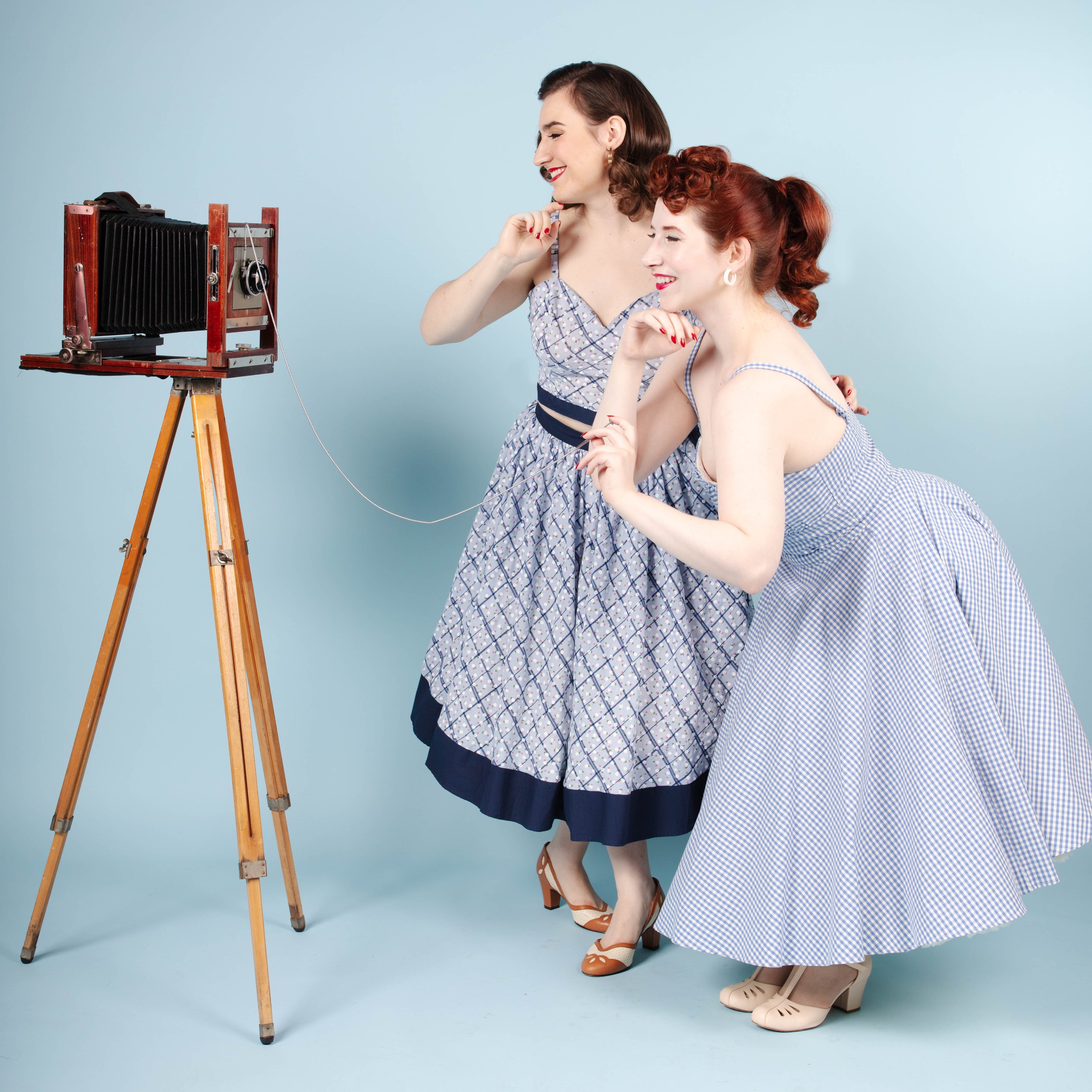 Two women dressed in vintage 1950s-style clothing, standing and crouching beside a large format camera on a tripod, smiling and looking at the camera.