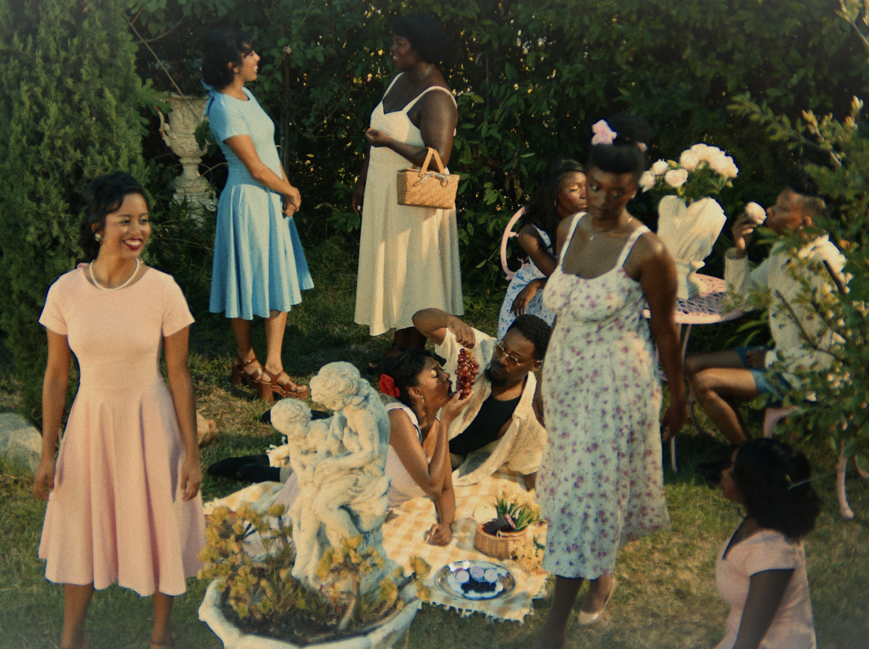 A group of women and children at a garden party, with some sitting on blankets on the grass and others standing, surrounded by greenery and flowers.