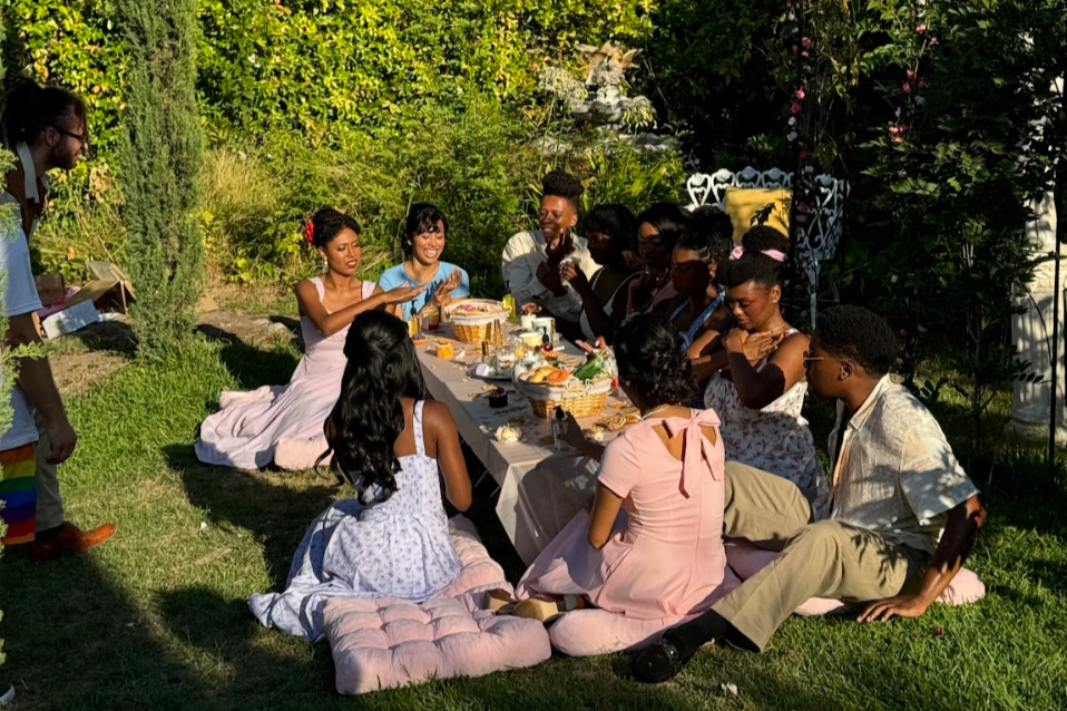 A group of people sitting around a table outdoors in a garden, having a meal or celebration. The setting is lush with greenery and flowers, and the attendees are engaged in conversation, with some smiling or laughing. There are baskets of food and dr