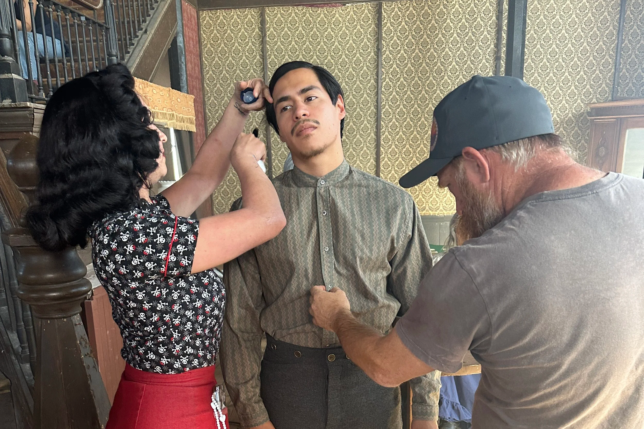 A young man with dark hair and a patterned shirt stands while two crew members, one woman with dark hair and a tattoo, and one man with a beard and a cap, are adjusting his makeup and costume in a vintage room with patterned wallpaper.