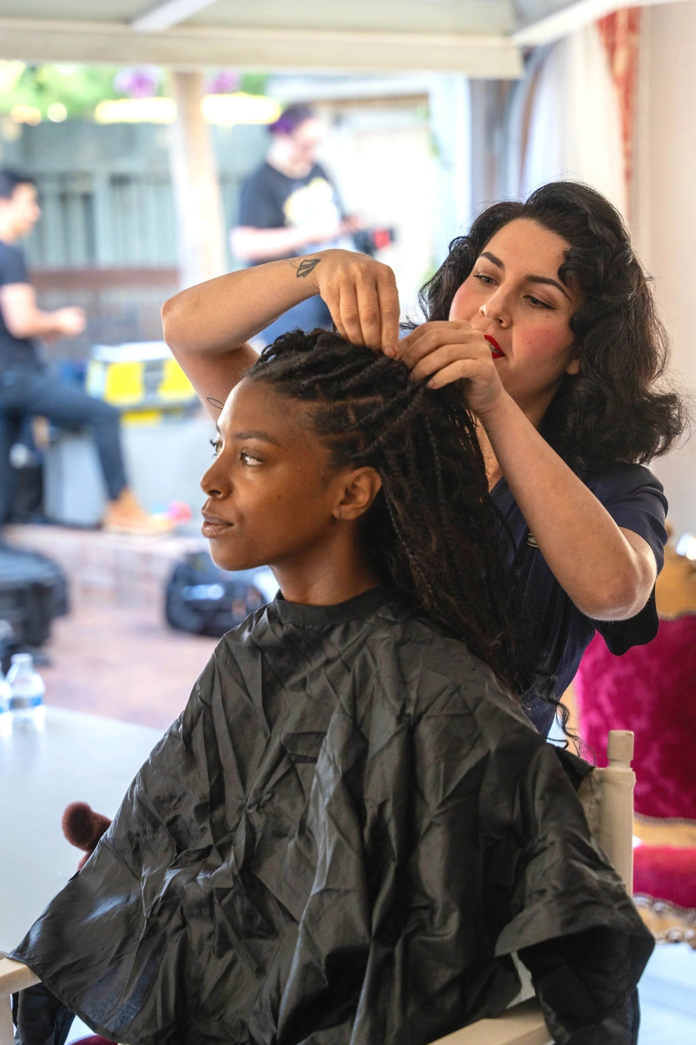 A woman with black hair is styling the hair of a seated woman with dreadlocks in a salon.