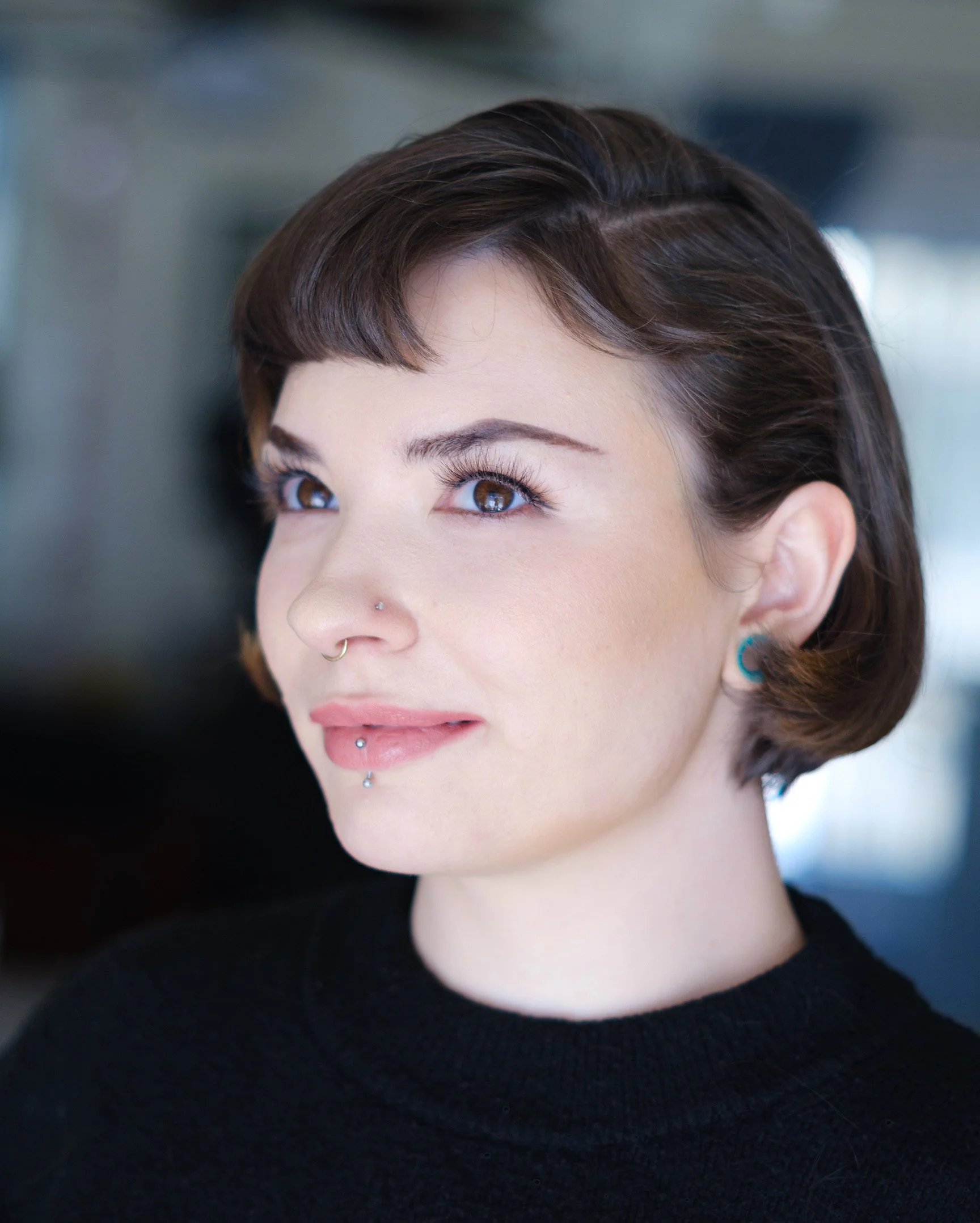 Close-up of a woman with short brown hair, blue eyes, wearing multiple facial piercings, and a black top, looking slightly upwards.