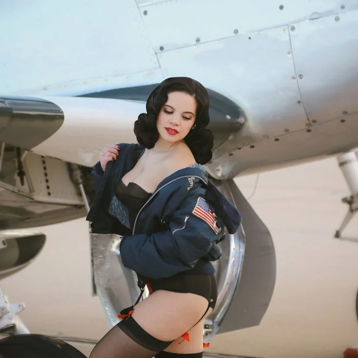 A woman in retro pin-up style outfit with a navy flight jacket and stockings posing next to a vintage aircraft, with part of the plane's engine and fuselage visible in the background.
