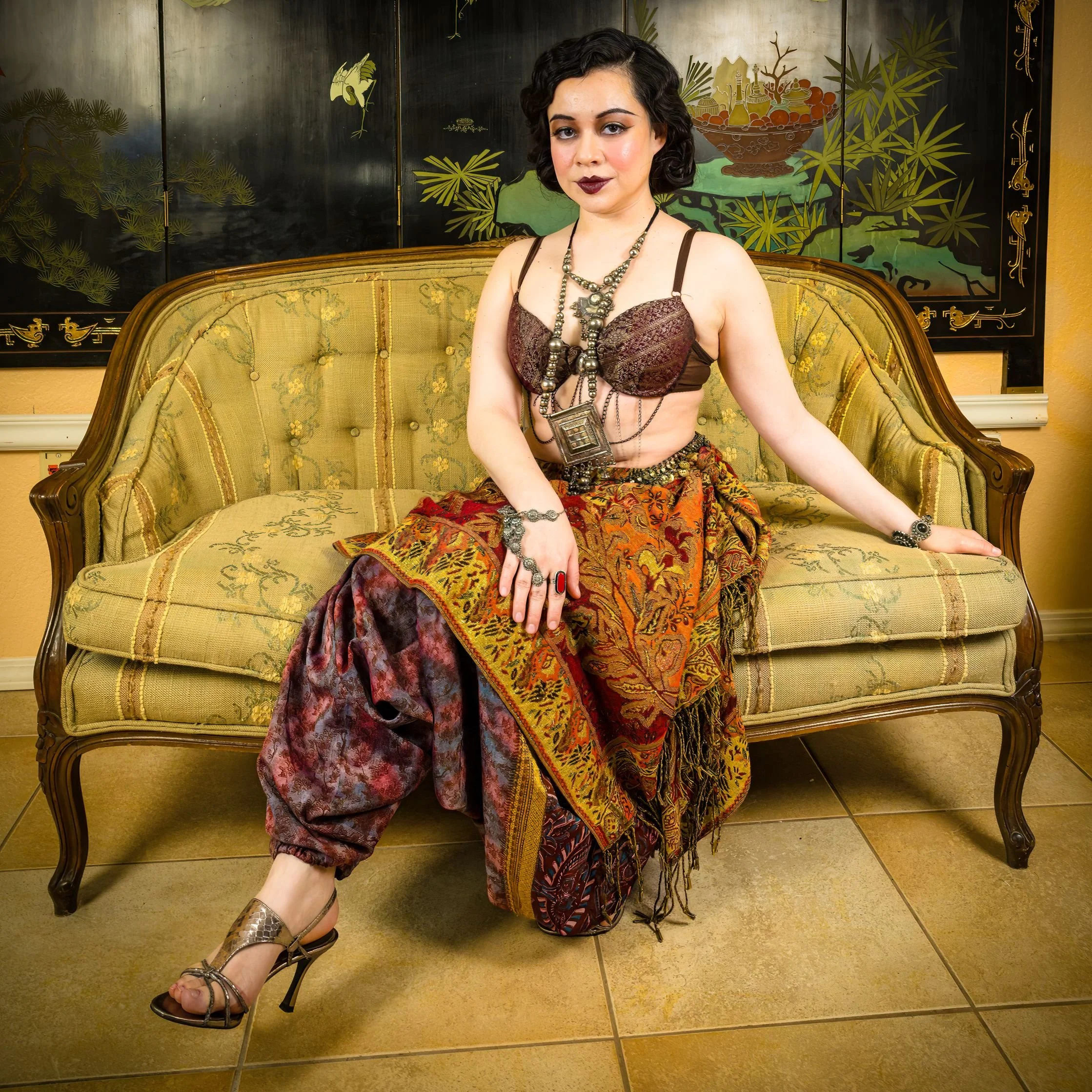A woman sitting on a vintage yellow and green sofa, dressed in an eclectic, bohemian style outfit with layered jewelry, dark lipstick, and patterned pants. She has curly black hair and is posed with one arm resting on the sofa.