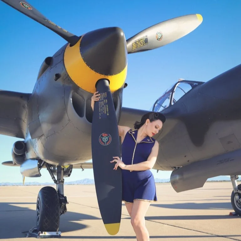 A woman with dark hair in a vintage style, wearing a blue outfit, stands next to a military aircraft on an airfield, holding one of the aircraft's propellers.