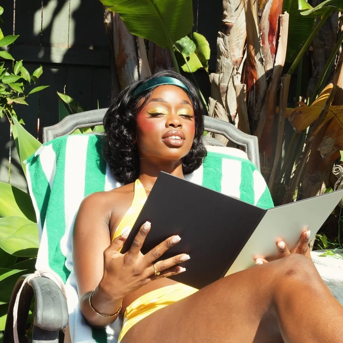 A woman with dark hair, wearing makeup, a headband, and yellow attire, is sitting outdoors on a garden chair with a green and white striped towel, reading a black book, surrounded by lush greenery and plants.