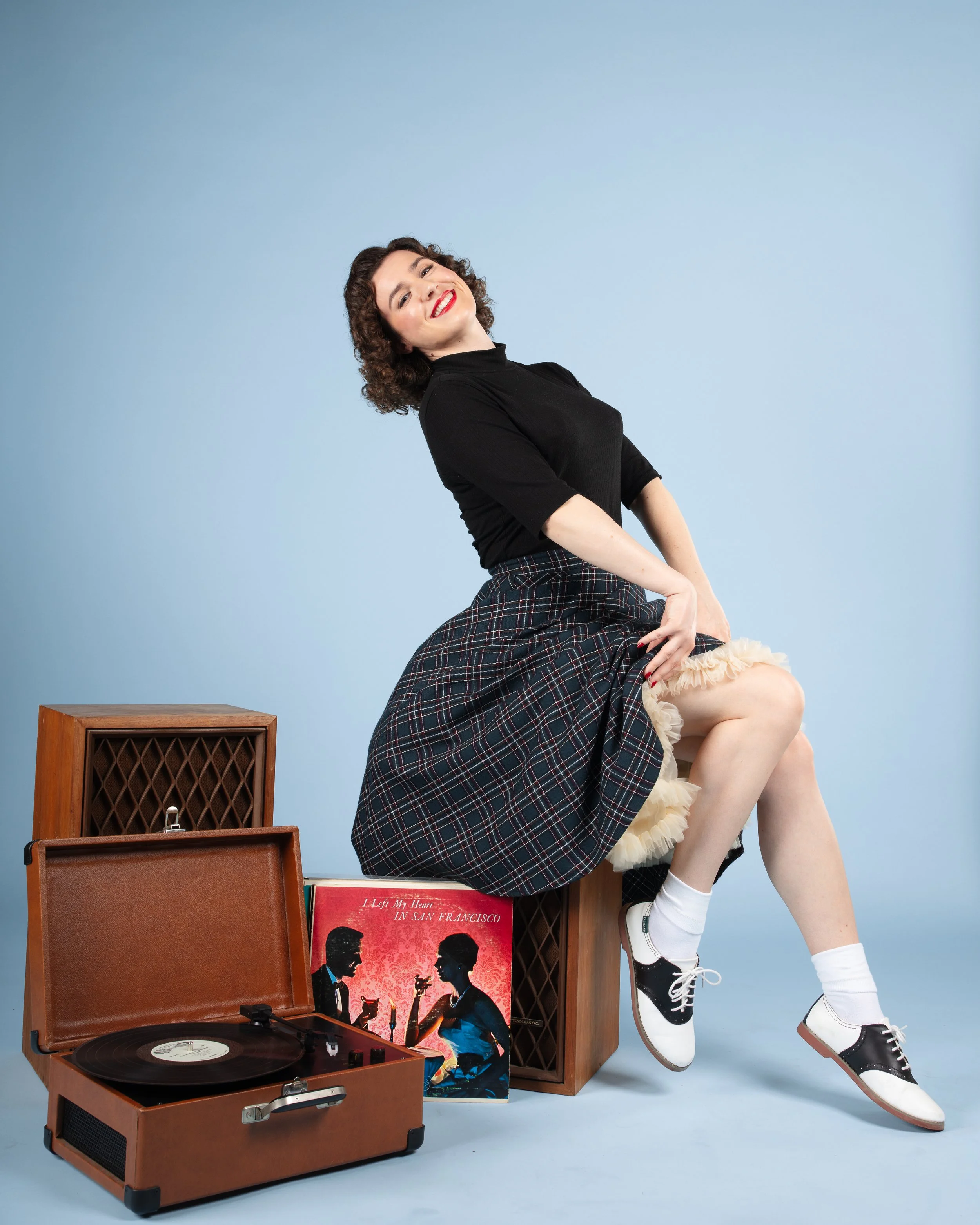 A woman with curly hair wearing a black top, plaid skirt, and white sneakers, smiling and sitting on a wooden speaker in front of a blue background. There is a vintage record player, a vinyl record, and a red album cover titled 'Lied My Heart in San Francisco' next to her.