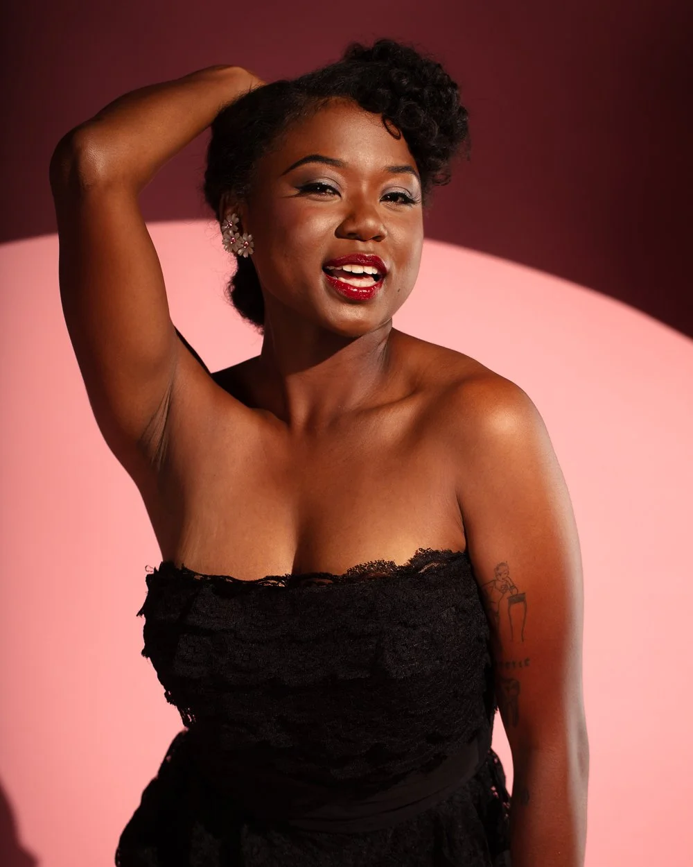 A woman with short curly hair, wearing a black strapless lace dress, smiling and posing with her right arm raised behind her head against a pink background.